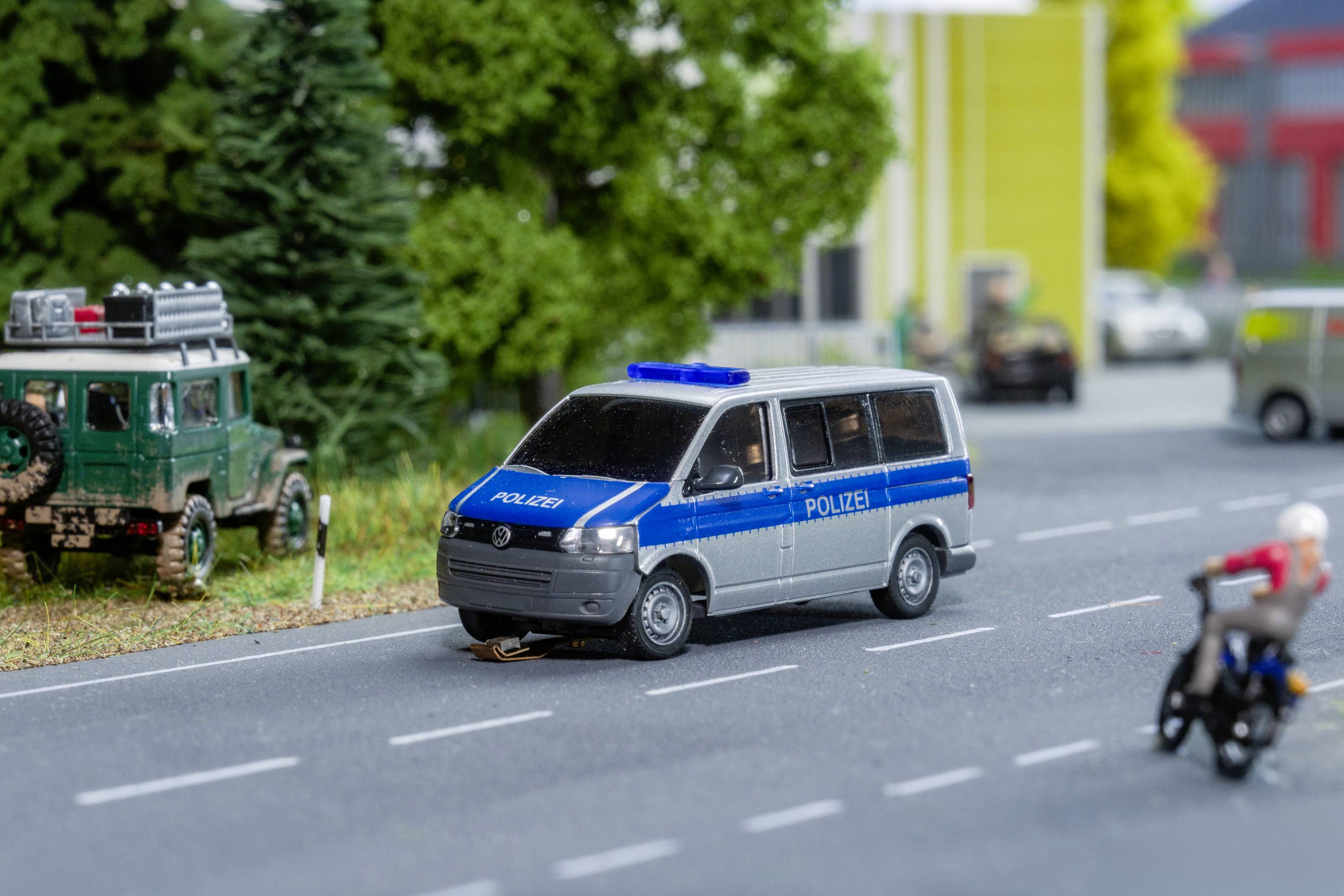 A blue and white police van is parked on a country road. In the background, a green off-road vehicle and blurry buildings can be seen.
