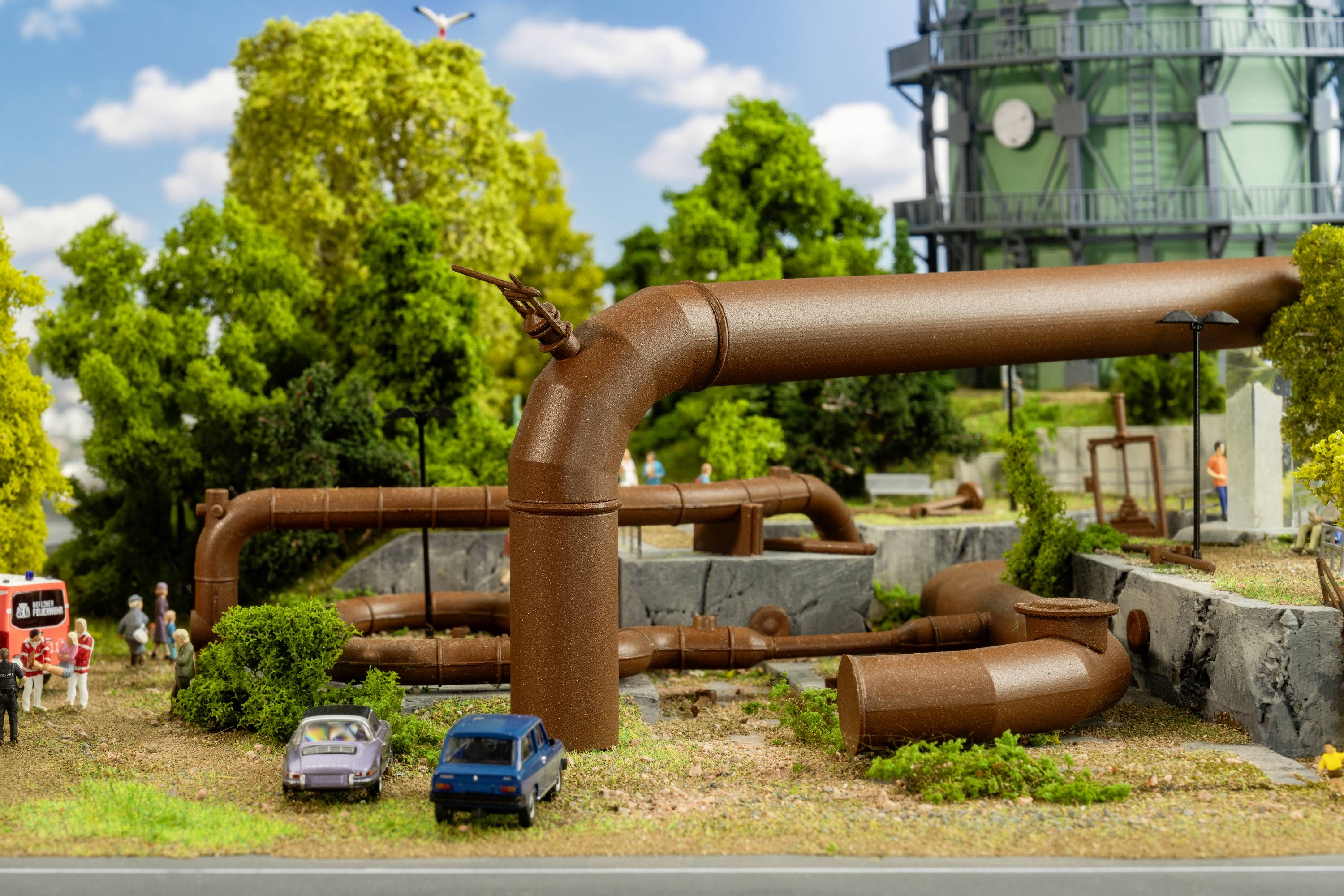 A miniature landscape with brown pipes in a green environment. Two cars are parked near the pipes, with a tower in the background.