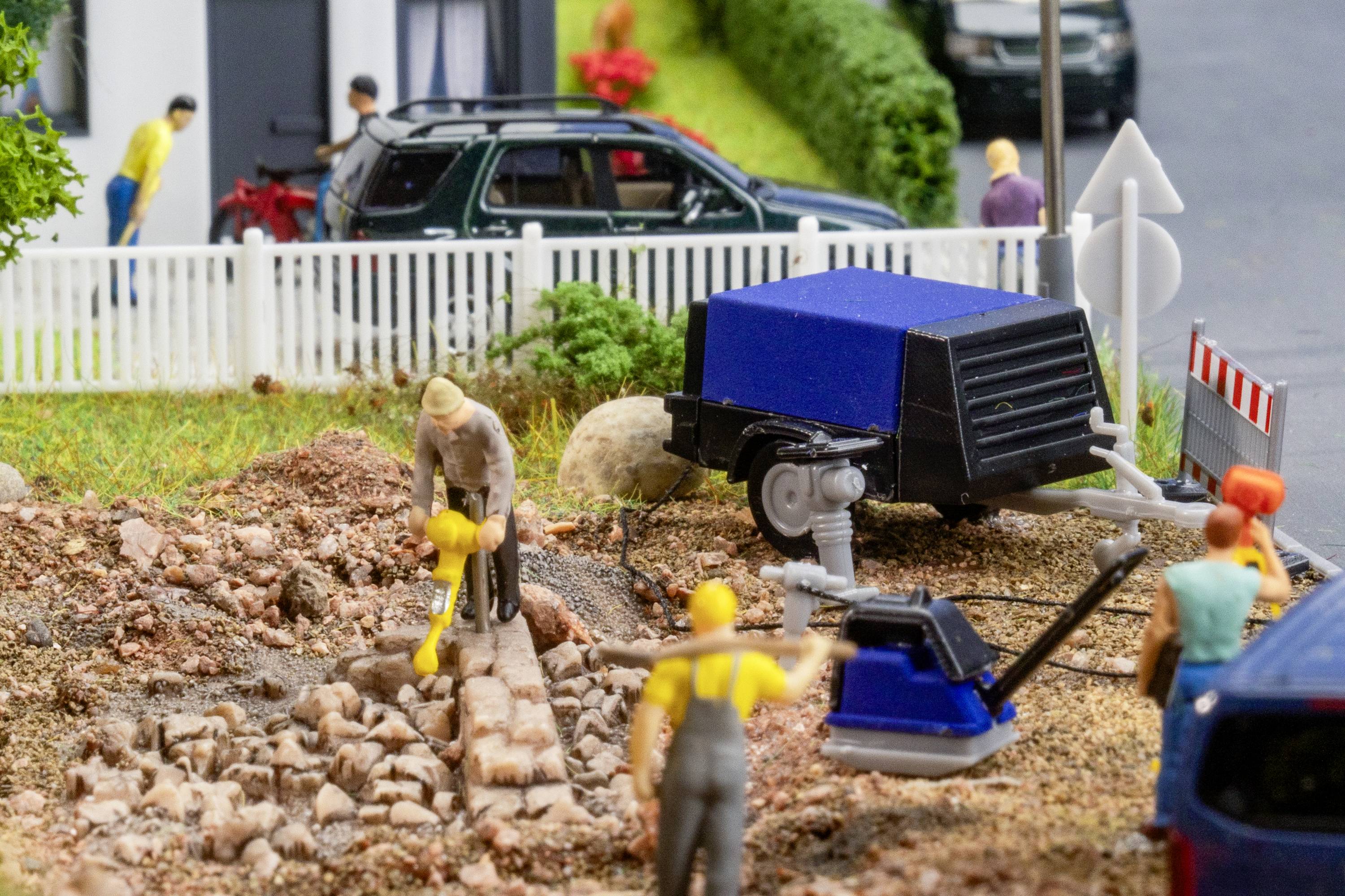Construction workers working on a road with machinery and barriers. In the background, a car is parked in front of a fence.