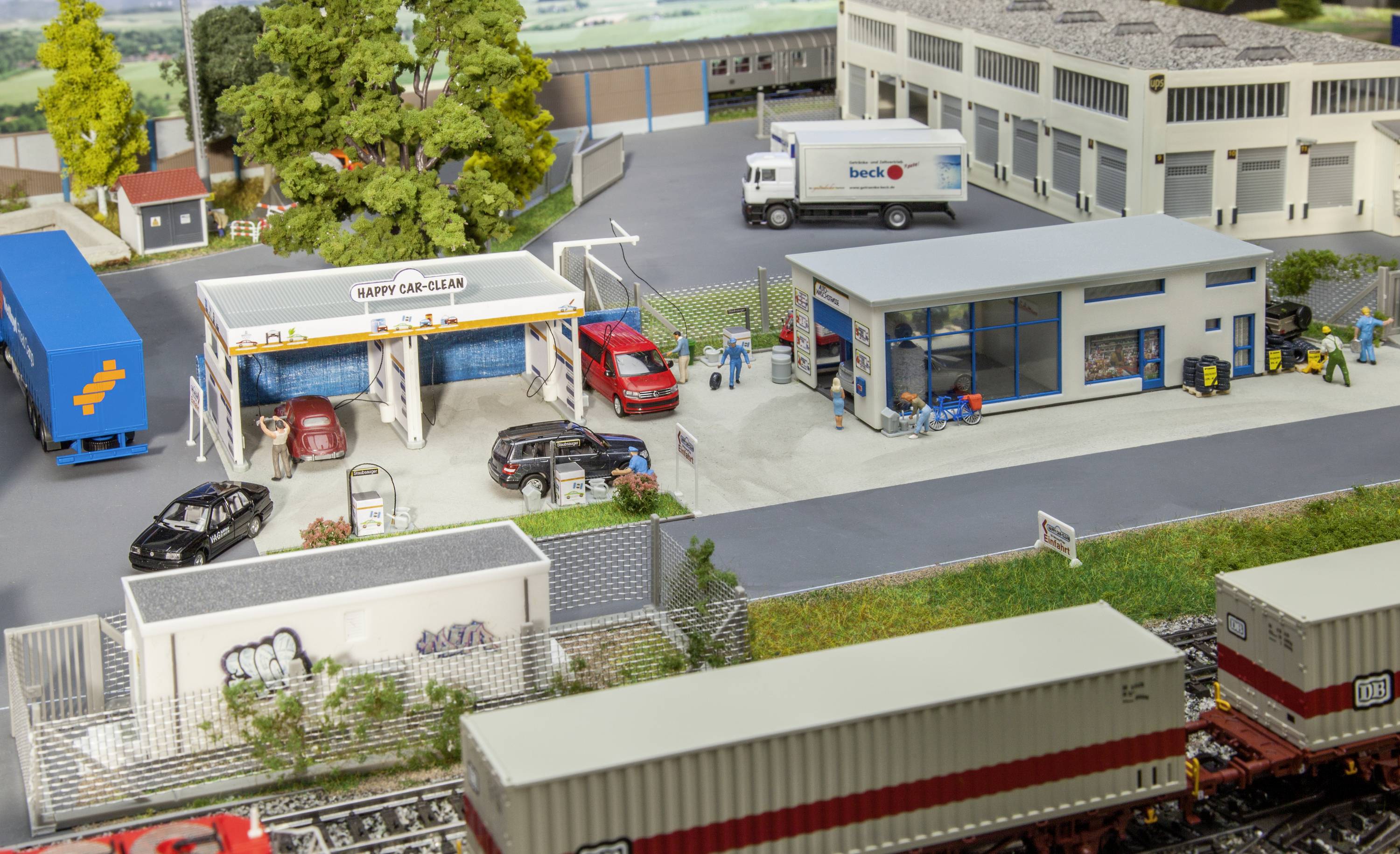 Miniature view of a car wash and workshop with cars and figures. Beside it, buildings, lorries and containers in a model landscape.