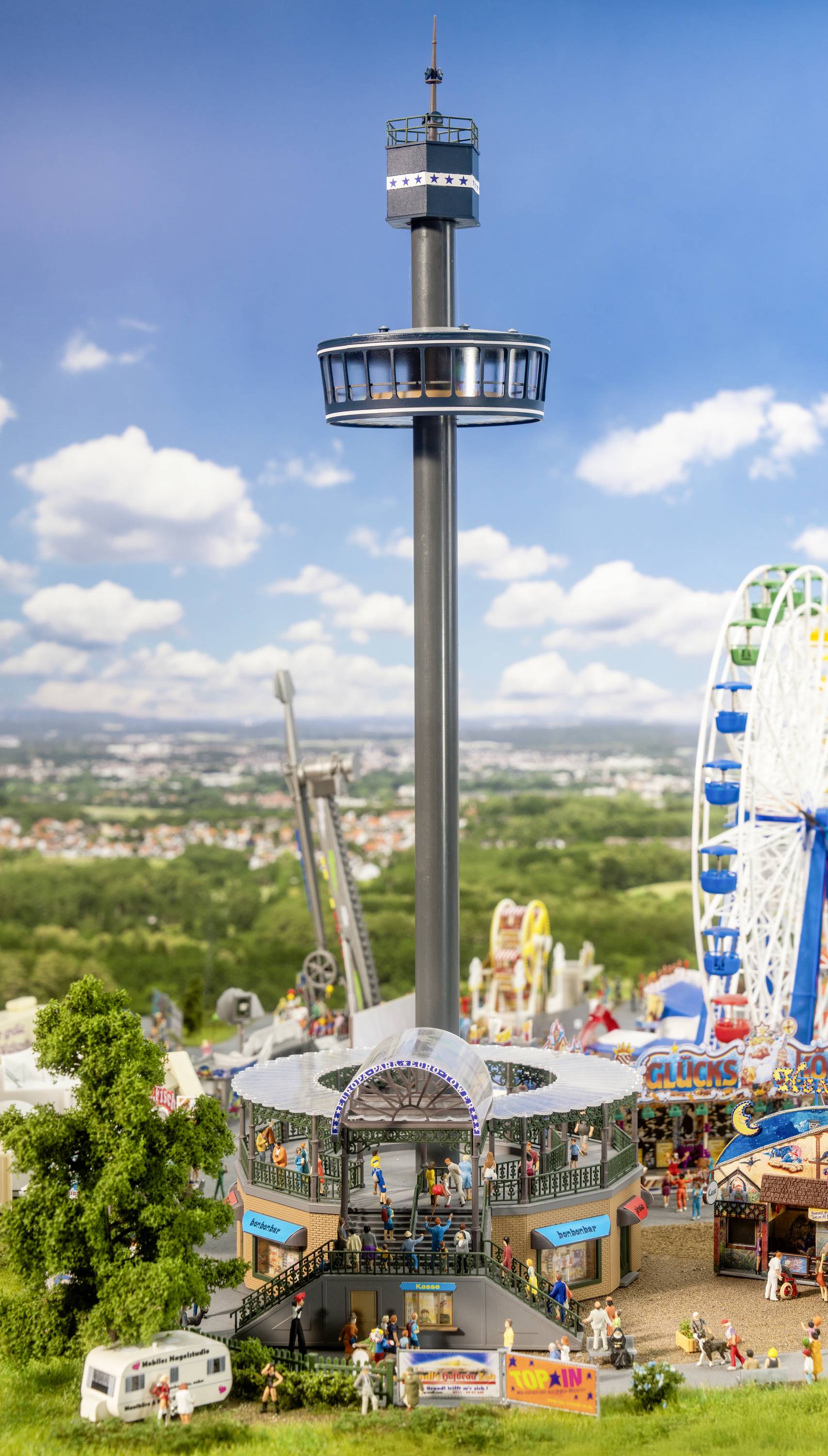 Observation tower at a fairground with people in the foreground, next to a Ferris wheel and other fairground rides; sunny day.