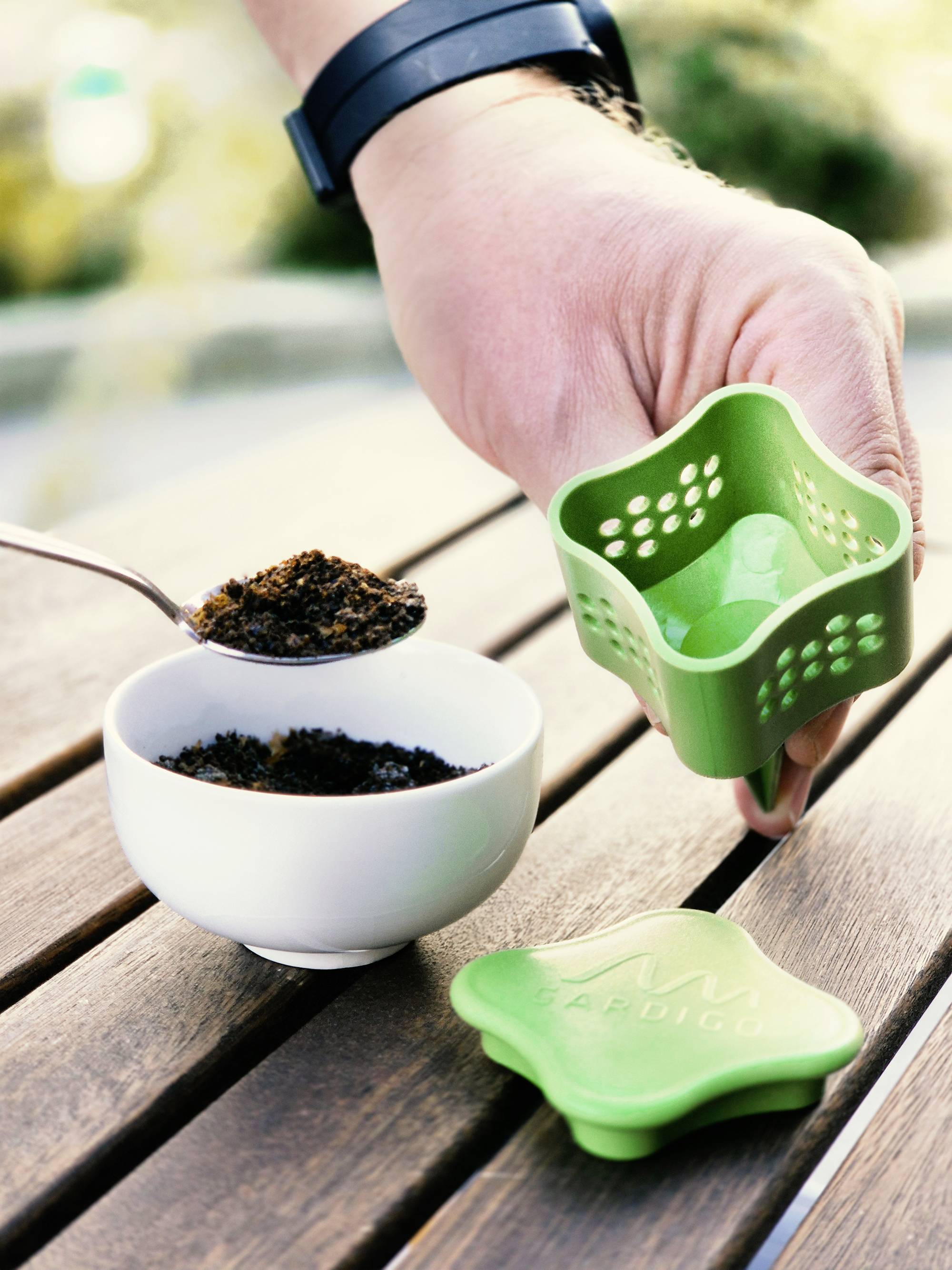 A hand is holding a green plastic container over a bowl of ground coffee on a wooden table. A spoon is being dipped into the bowl.