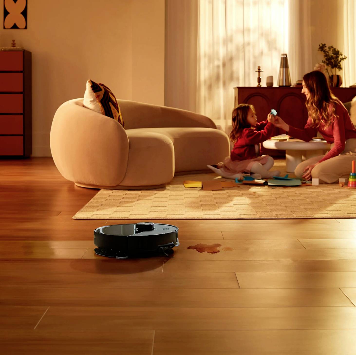 A robotic vacuum cleaner cleans the floor, while in the background a woman plays with a child at the table.