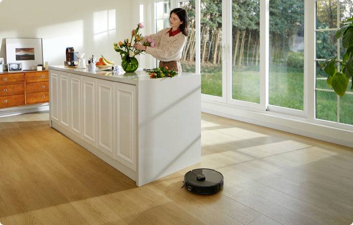 A person is arranging flowers on a kitchen worktop, while a vacuum robot drives across the wooden floor in the foreground.