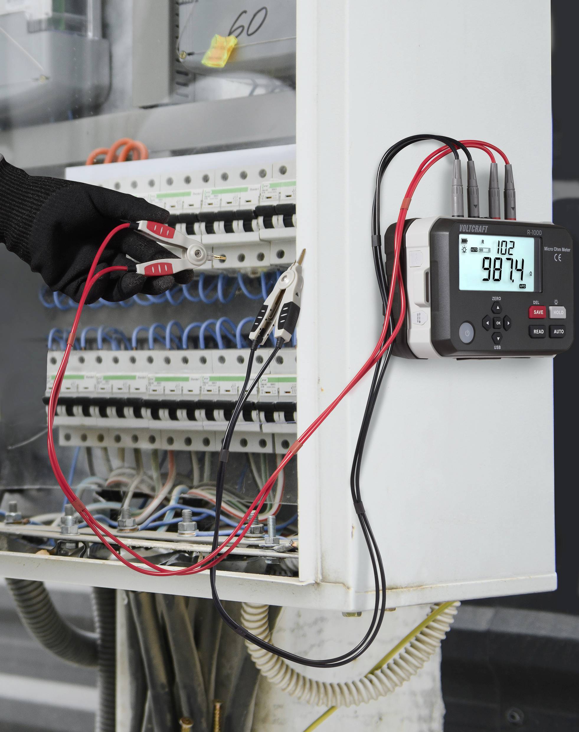 An engineer's gloved hand uses a red clamp meter to measure current in an electrical panel, displaying '98.74' on its digital screen.