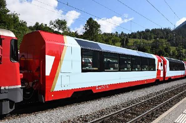 A red and white train travels on tracks through a green, hilly landscape under a blue sky with few clouds.