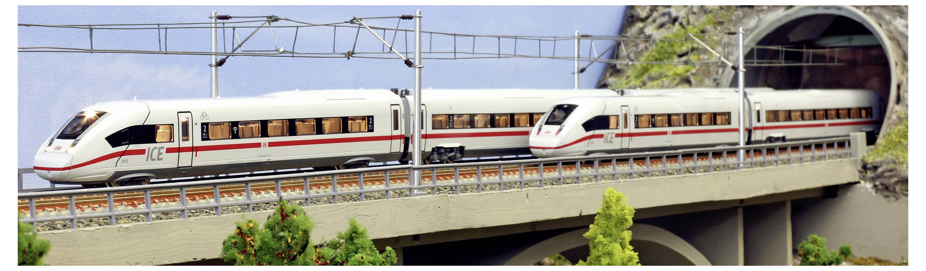 A model train, ICE-like, exits a tunnel on an elevated railway track through a landscape with trees in the foreground.