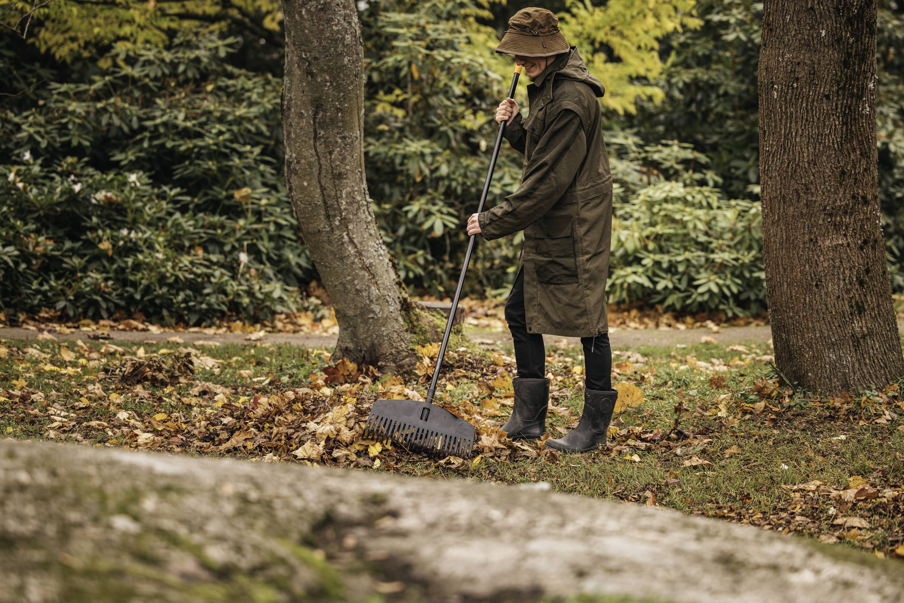 A person wearing green clothing and wellington boots is raking leaves in an autumnal park. Trees and piles of leaves are visible in the background.