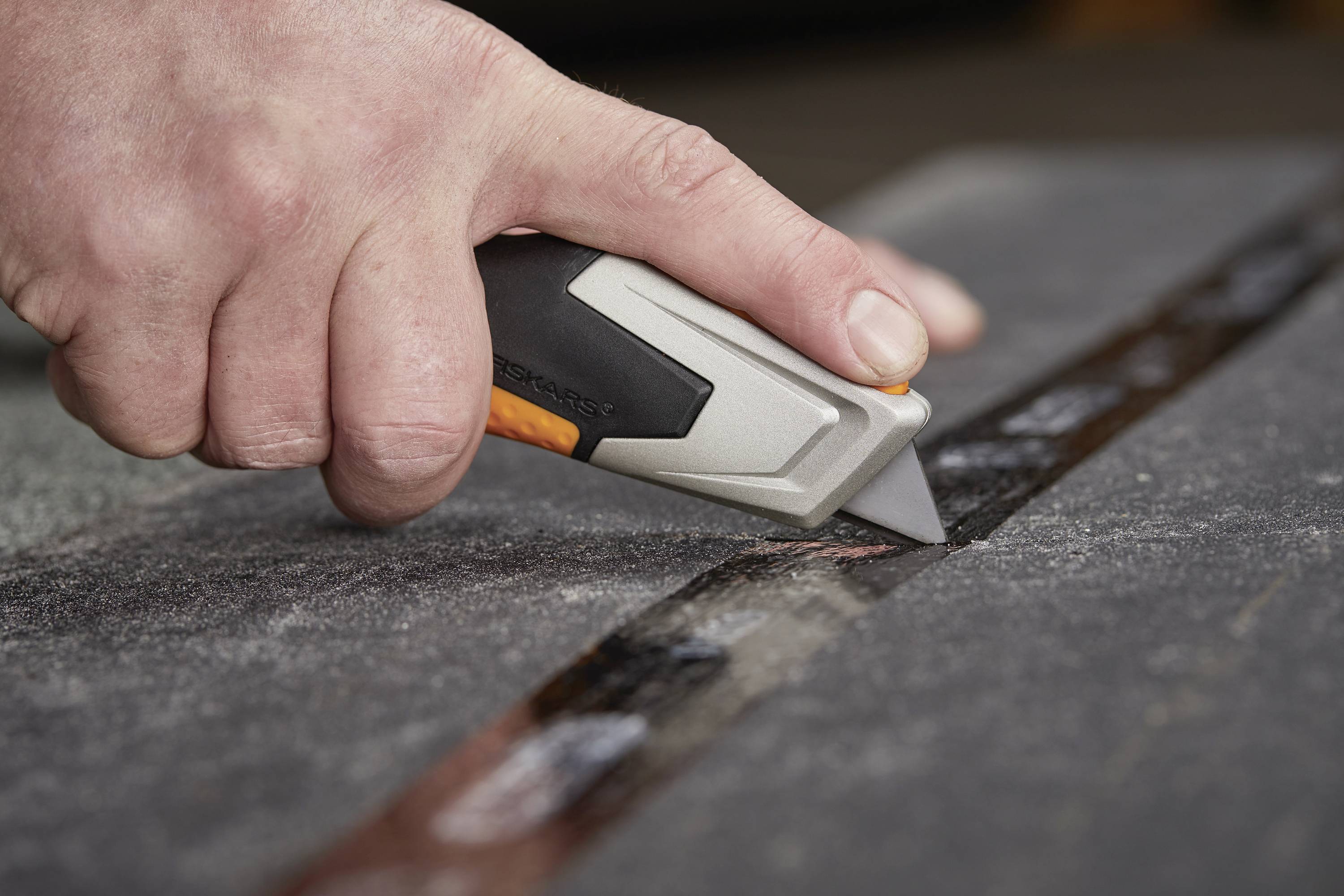 A hand holds a craft knife and cuts a straight line into a dark fabric or carpet.