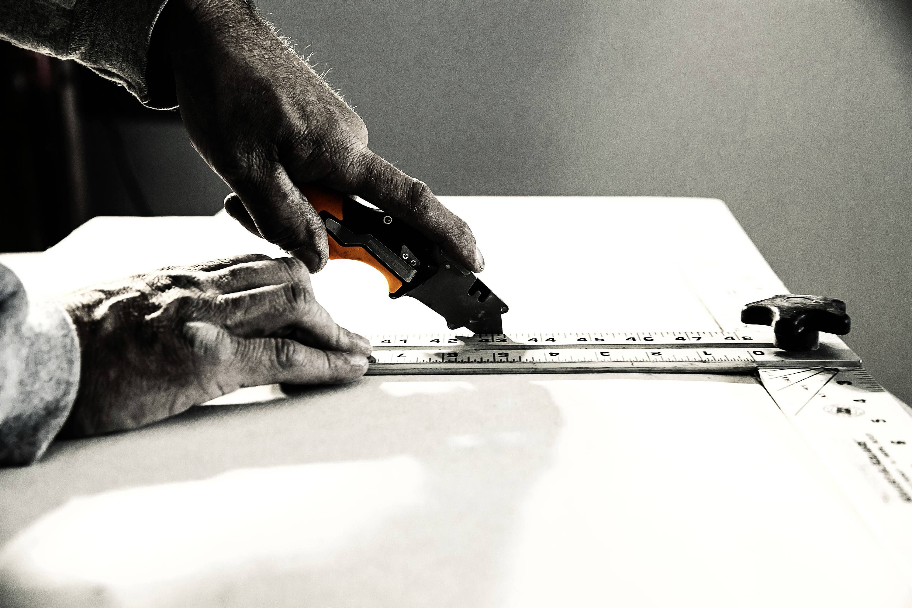A person is cutting a piece of material along a metal ruler using a craft knife. Hands are strongly focused.