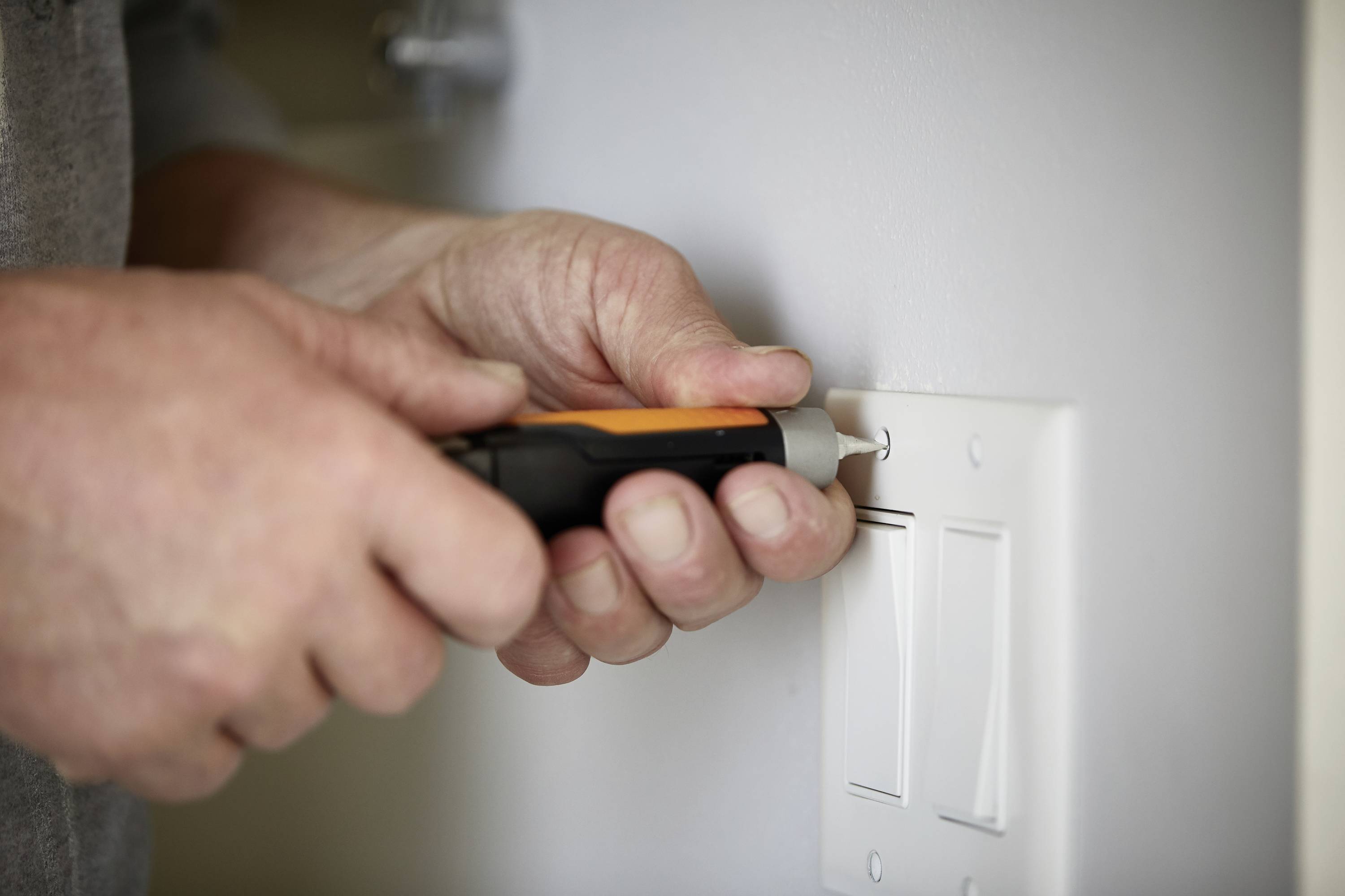 A person is installing or repairing a light switch with a screwdriver on a white wall.