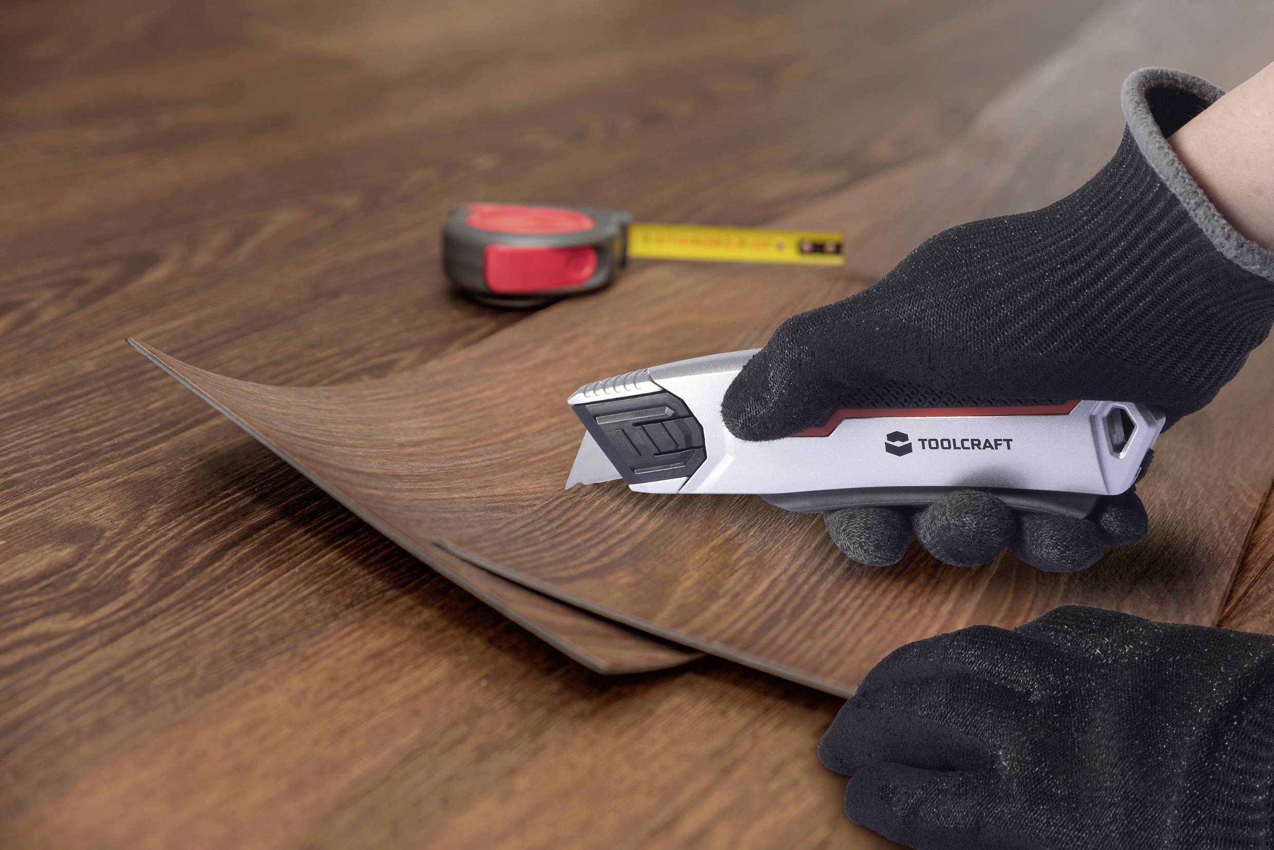 A gloved hand uses a utility knife to cut a piece of wood-patterned laminate on a table. A tape measure is in the background.