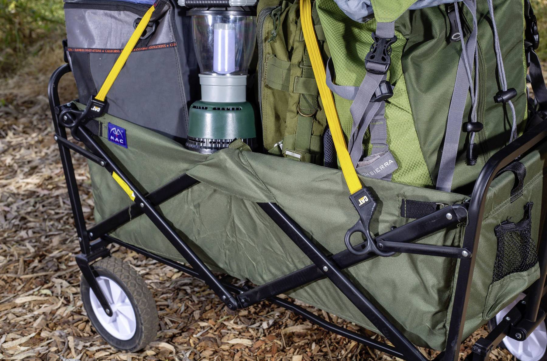 A green folding wagon filled with camping gear, including backpacks, a lantern, and a cooler, on a leaf-covered forest floor.