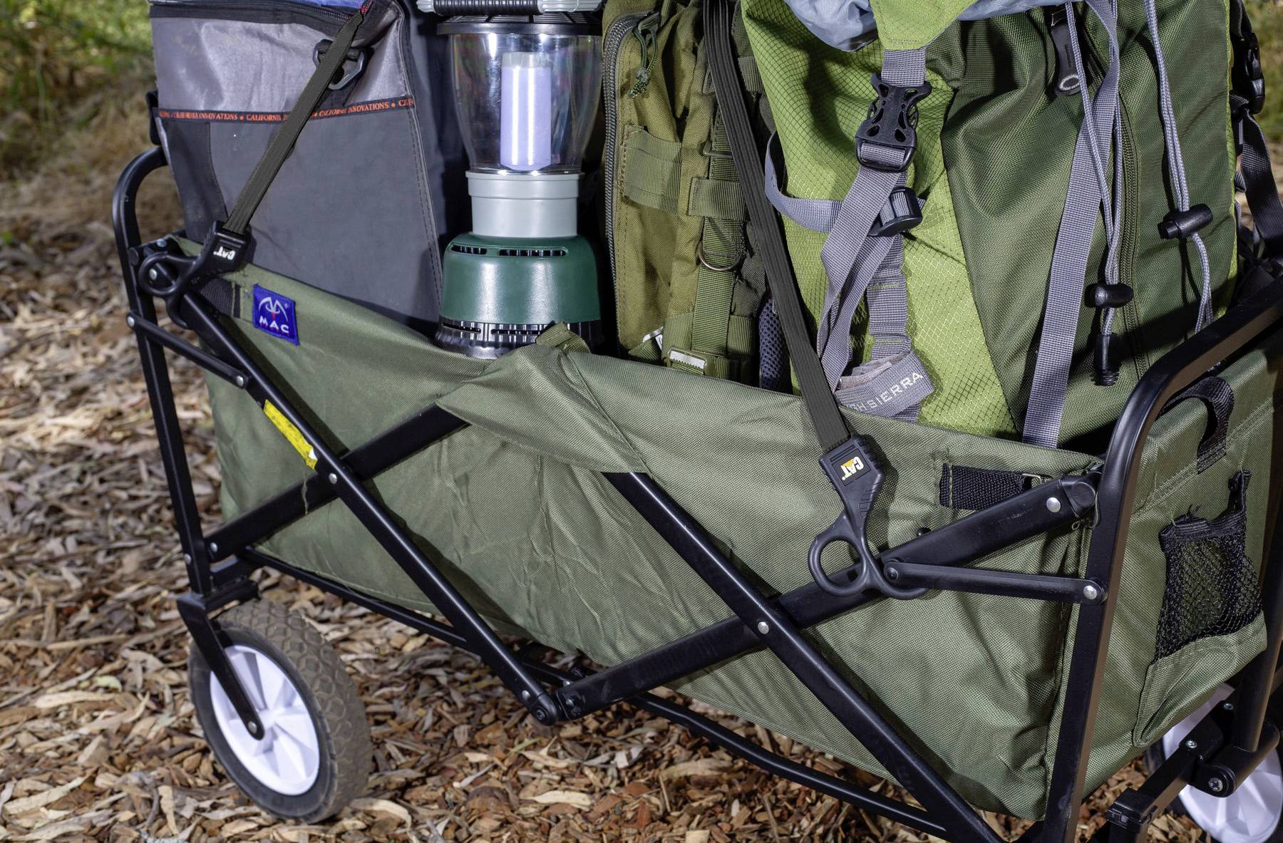 A green folding utility wagon carrying hiking backpacks, a lantern, and outdoor equipment, set on a trail covered with fallen leaves.