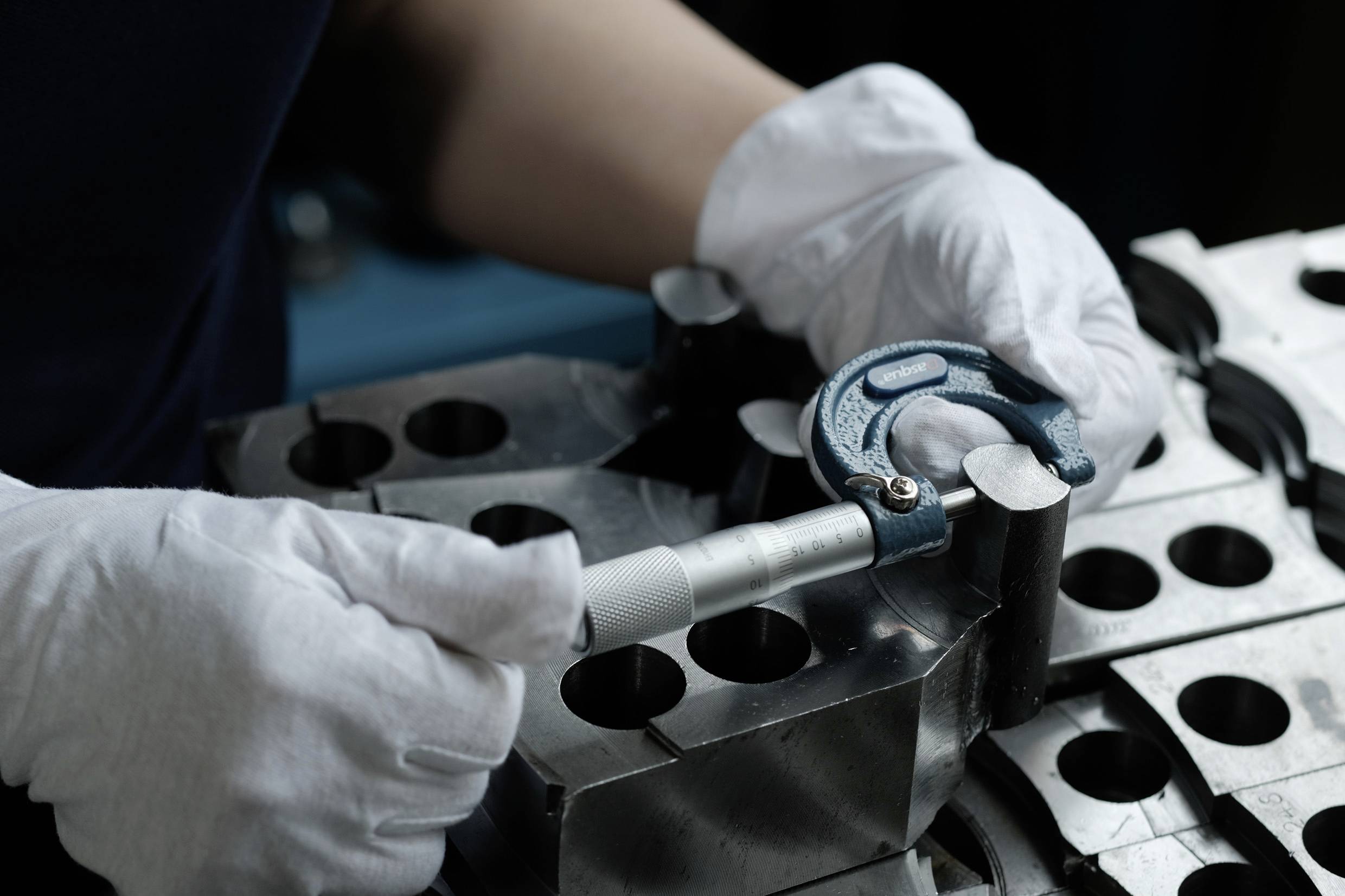 A person wearing white gloves measures a metal block using a micrometer gauge, focusing on precision and accuracy in a workshop setting.