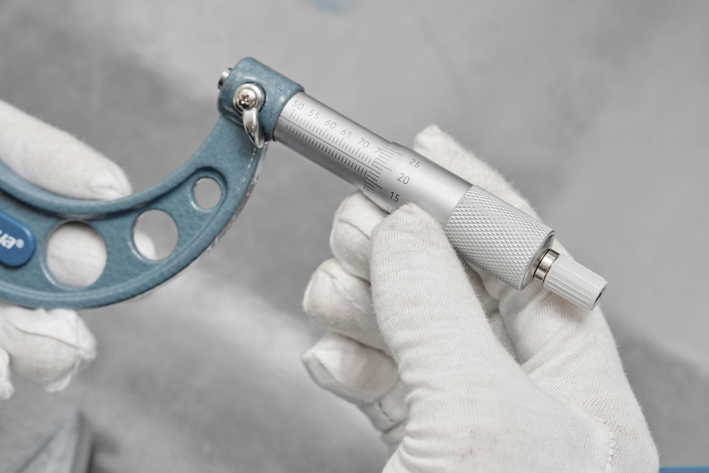 A person wearing white gloves is holding a micrometer, measuring a small object's dimensions with precision against a gray background.