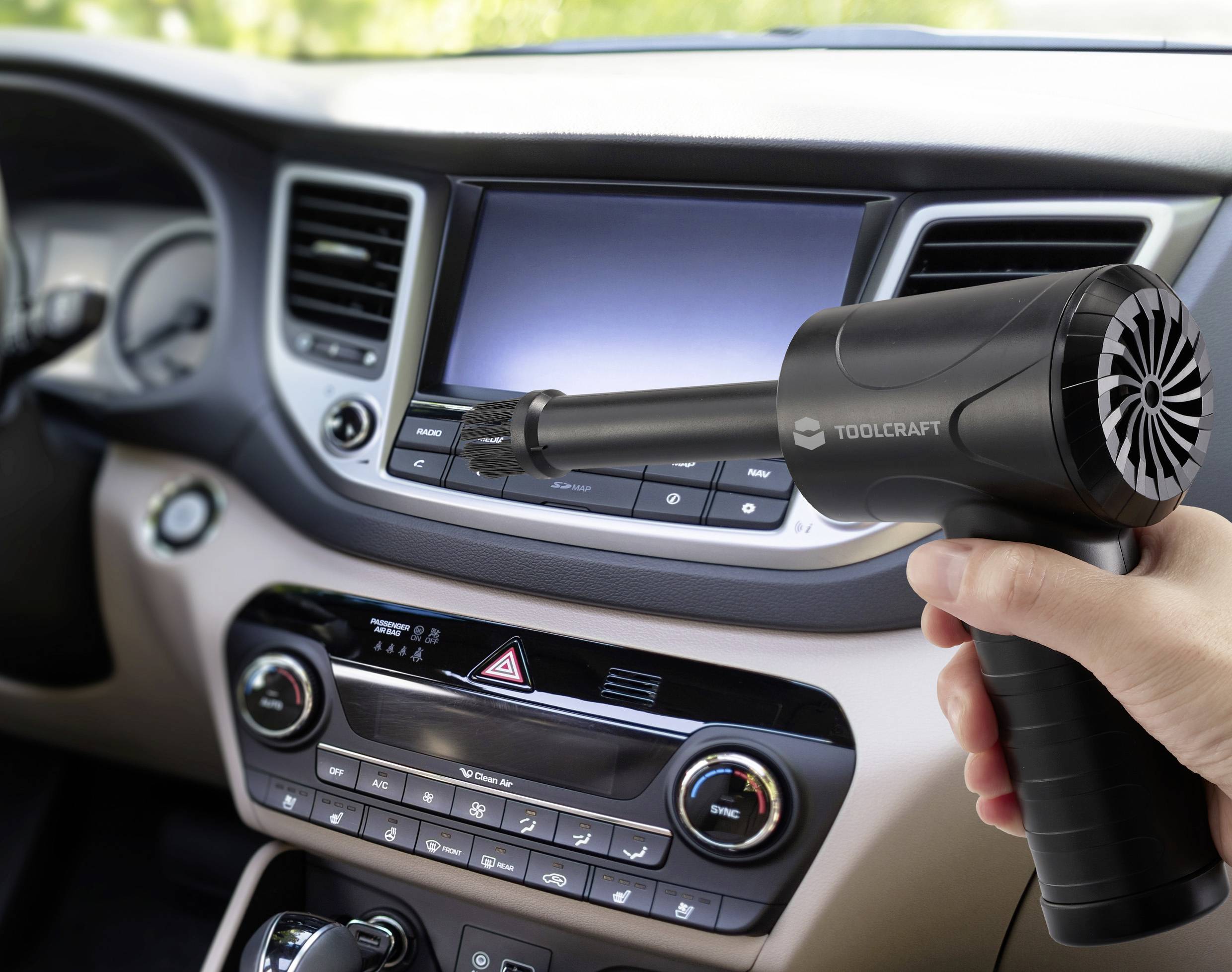 A person holds a small black air blower labeled 'Toolcraft' inside a car, aimed at cleaning the area around the dashboard and controls.