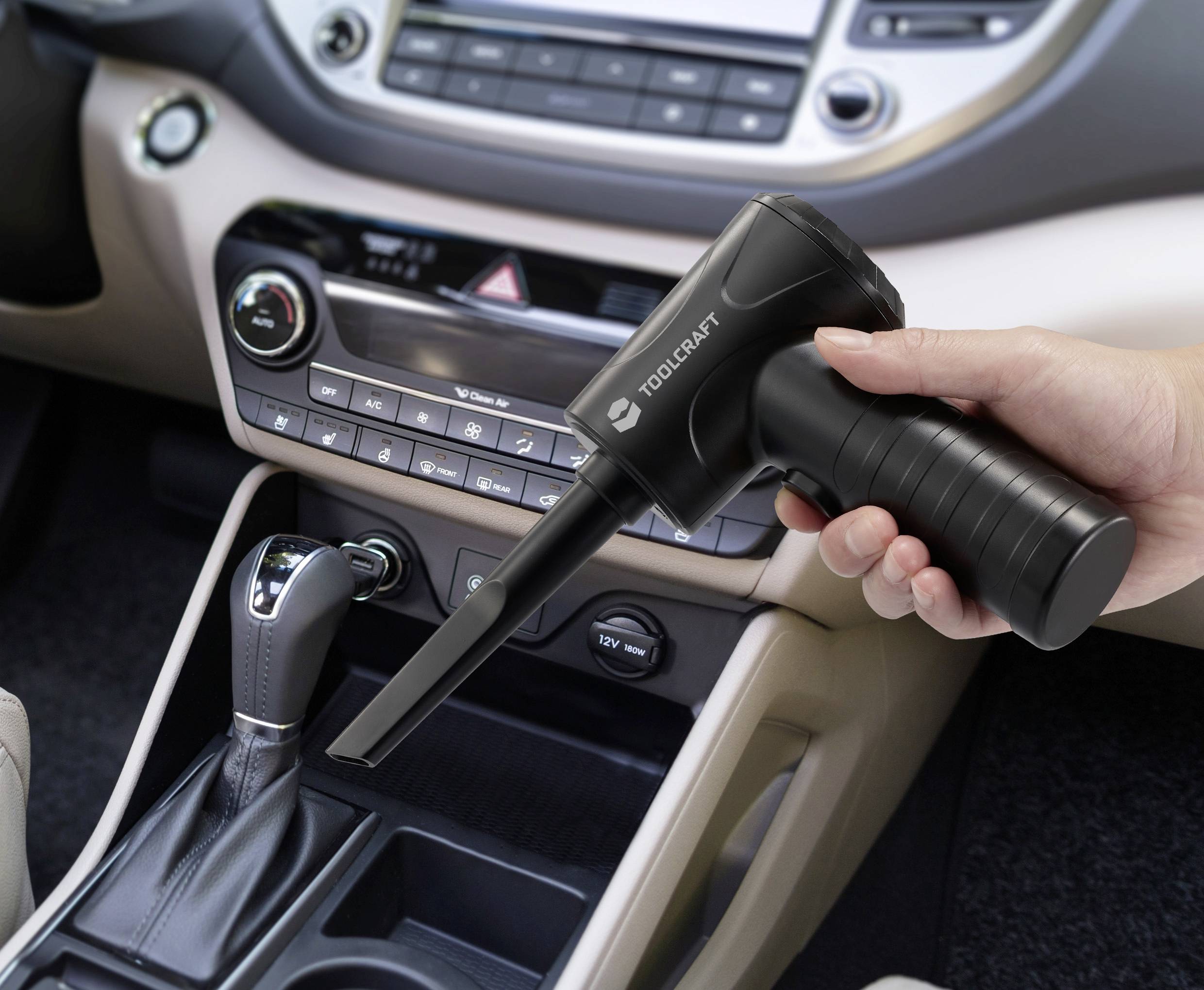 Hand holding a compact black air duster in a car interior, aimed at the gear shift area for cleaning debris.
