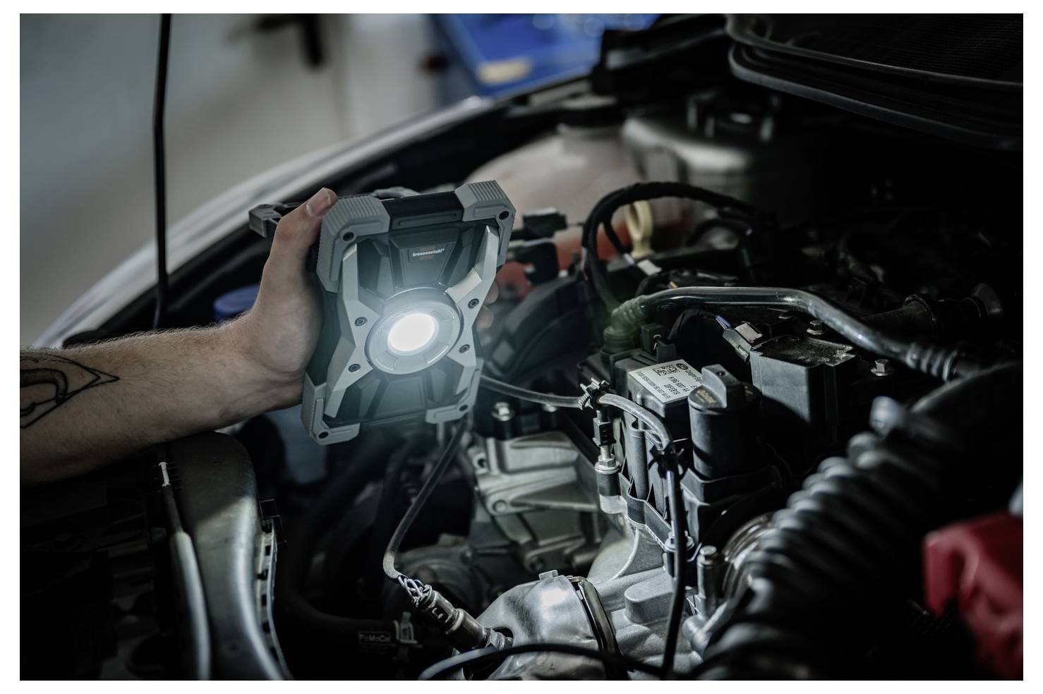A person holds a lit work light inside a car engine bay, inspecting or repairing the engine