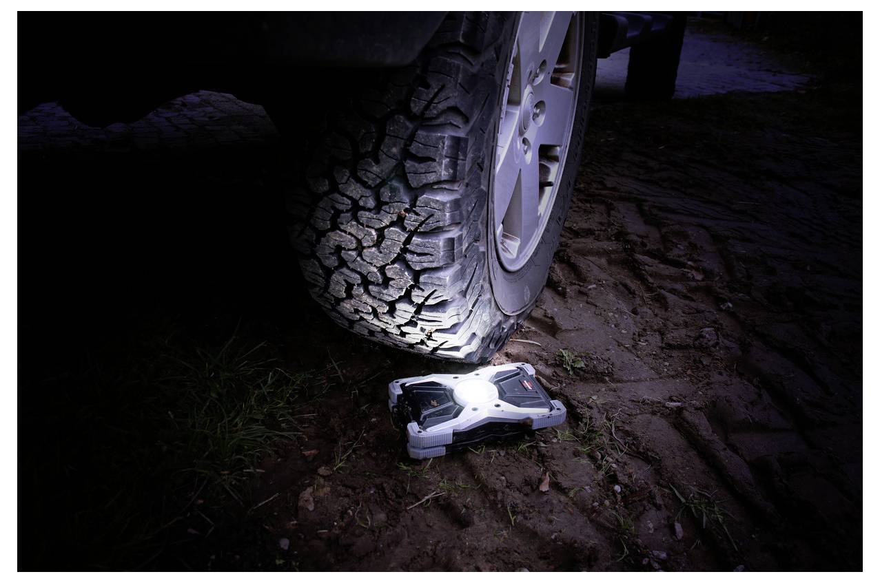 A car tire is pressing on a small rugged remote-controlled vehicle on muddy ground at night, crushing it under the wheel.