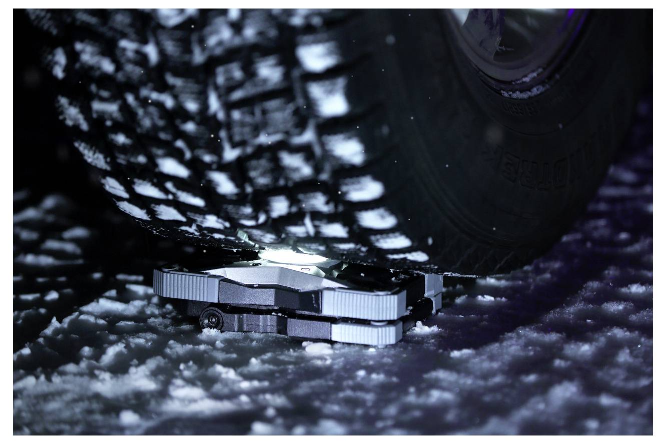 Close-up of a vehicle wheel and undercarriage resting on snow or ice, highlighting a compact metal traction device beneath the tire.