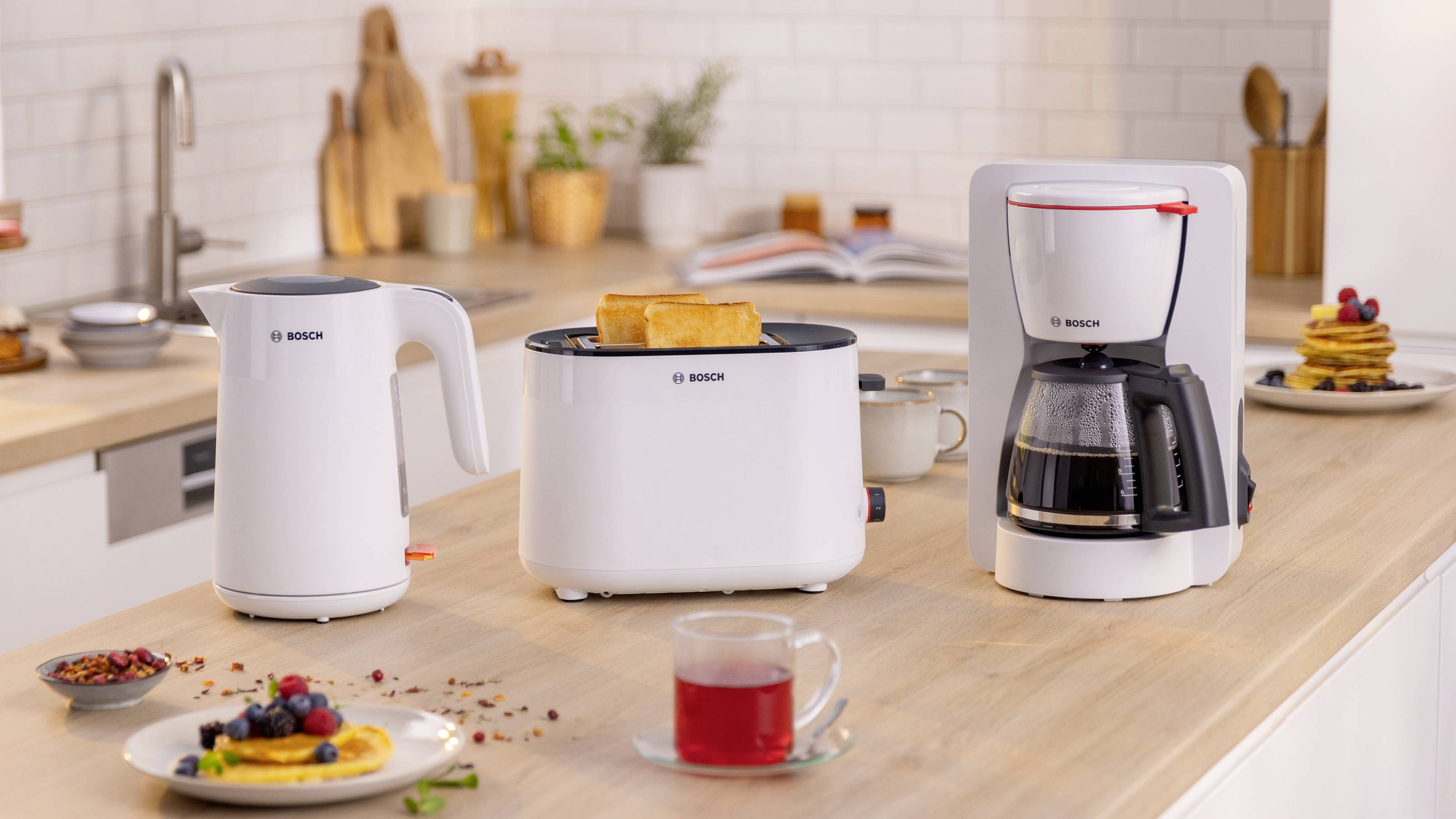 A modern kitchen counter with a white electric kettle, toaster with toast slices, and coffee maker, surrounded by breakfast items.