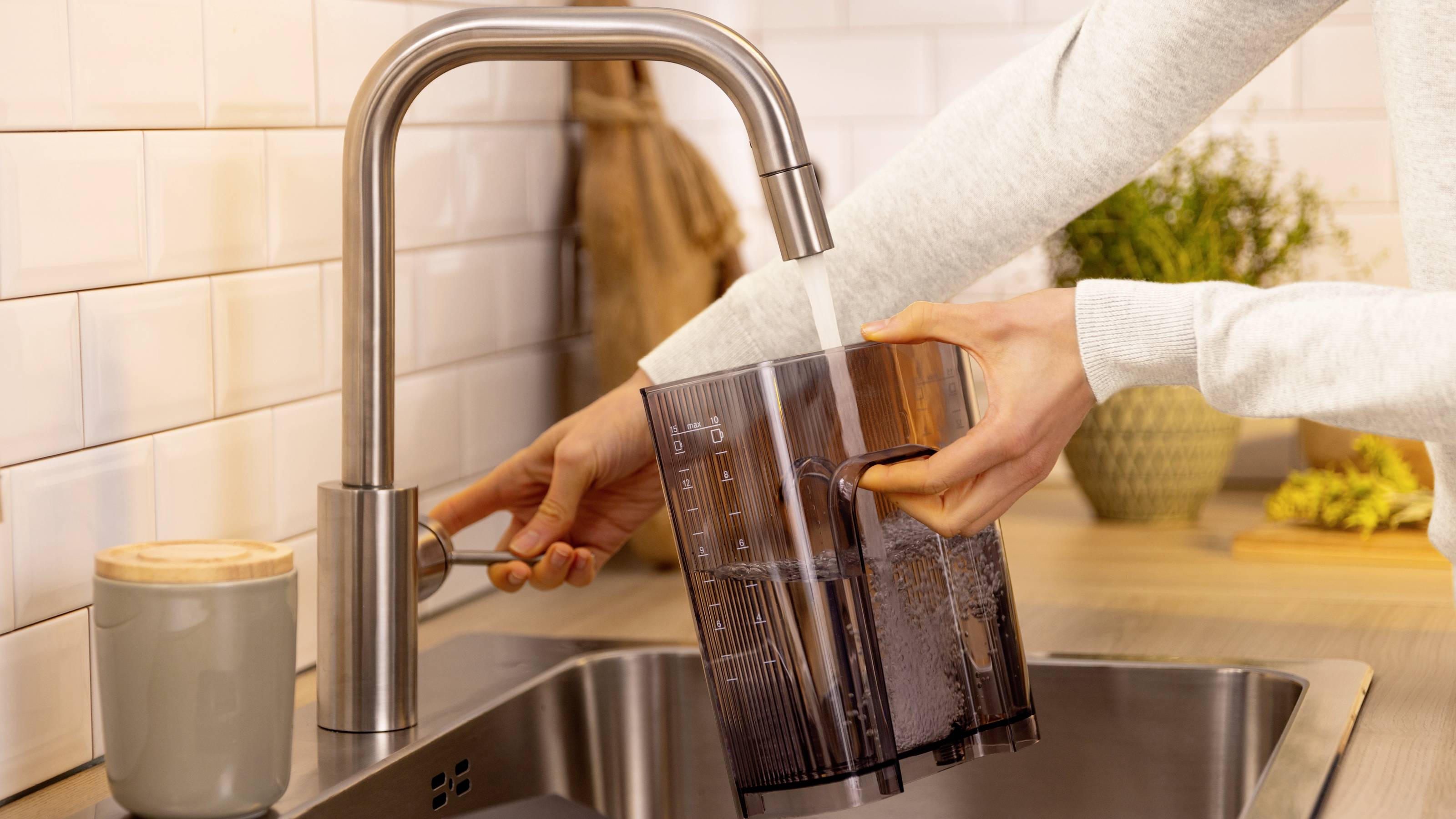 A person fills a transparent coffee pot with water from a modern kitchen faucet, while a gray mug sits on the counter nearby.