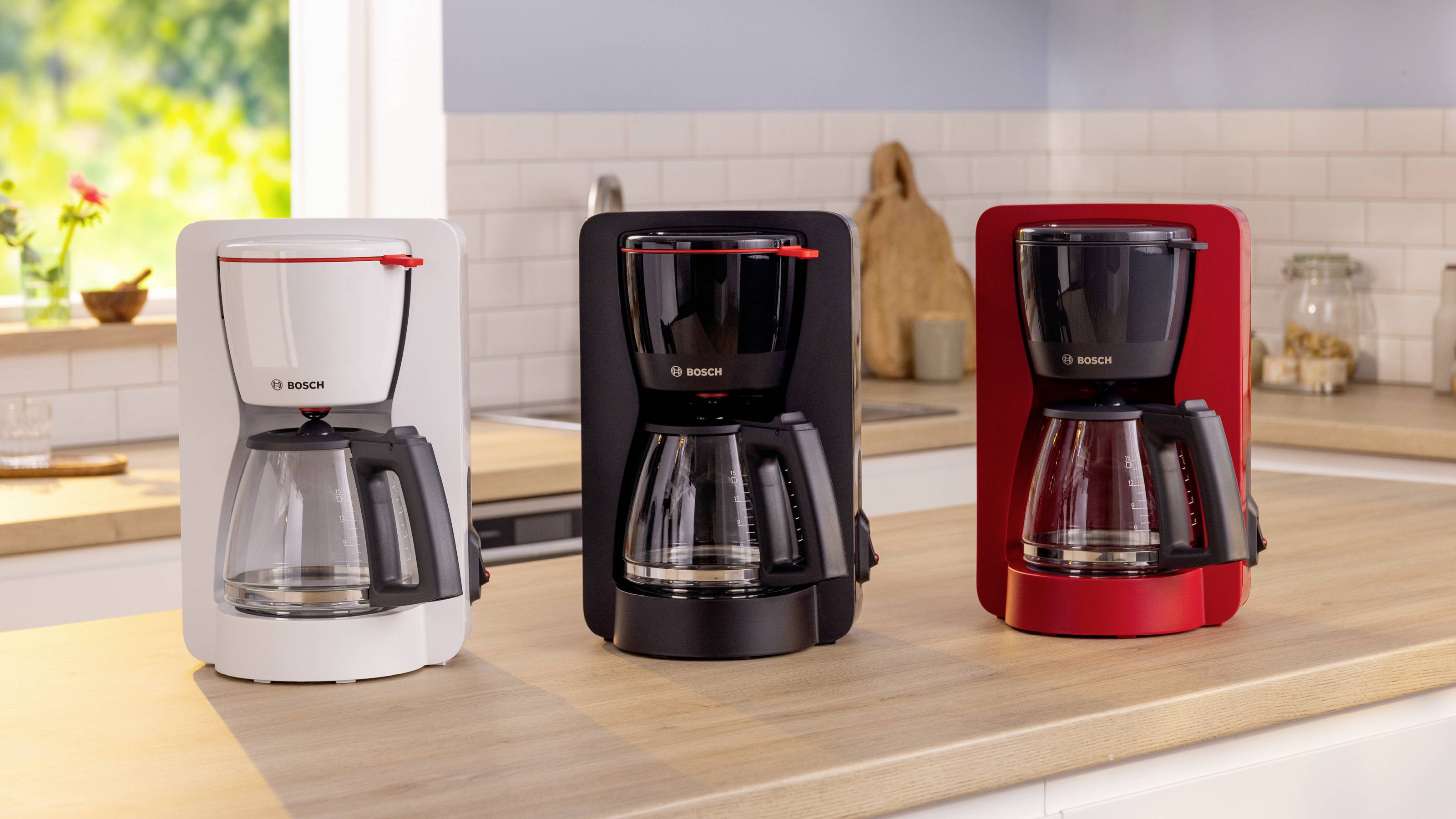 Three coffee makers in white, black, and red are placed on a kitchen counter with a tiled backsplash and blurred background.