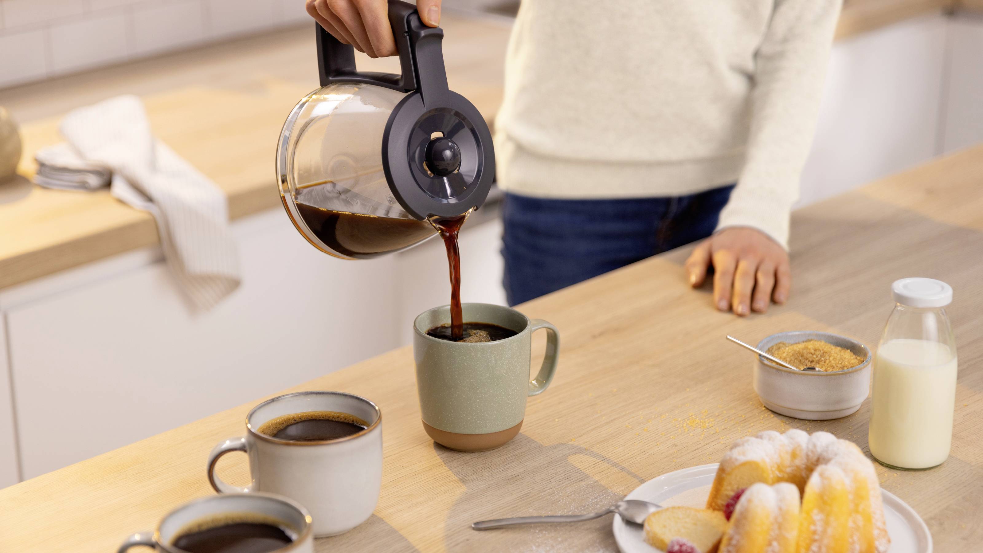 A person pours coffee from a pot into a mug on a kitchen counter, surrounded by two other mugs, a plate of pastries, sugar, and milk.