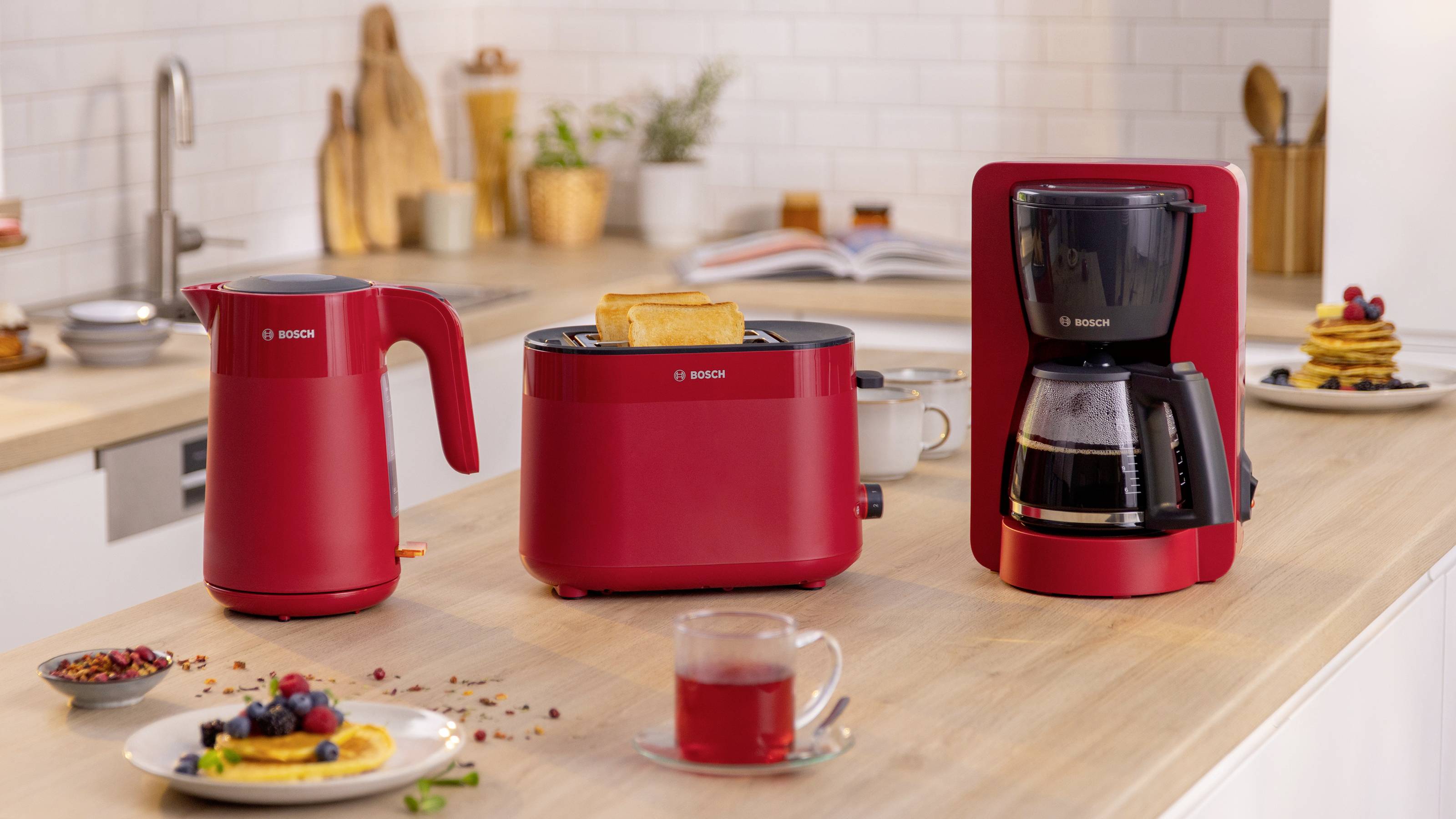 A kitchen countertop features a red electric kettle, toaster with toast, and coffee maker. In the background are pancakes and fruit.