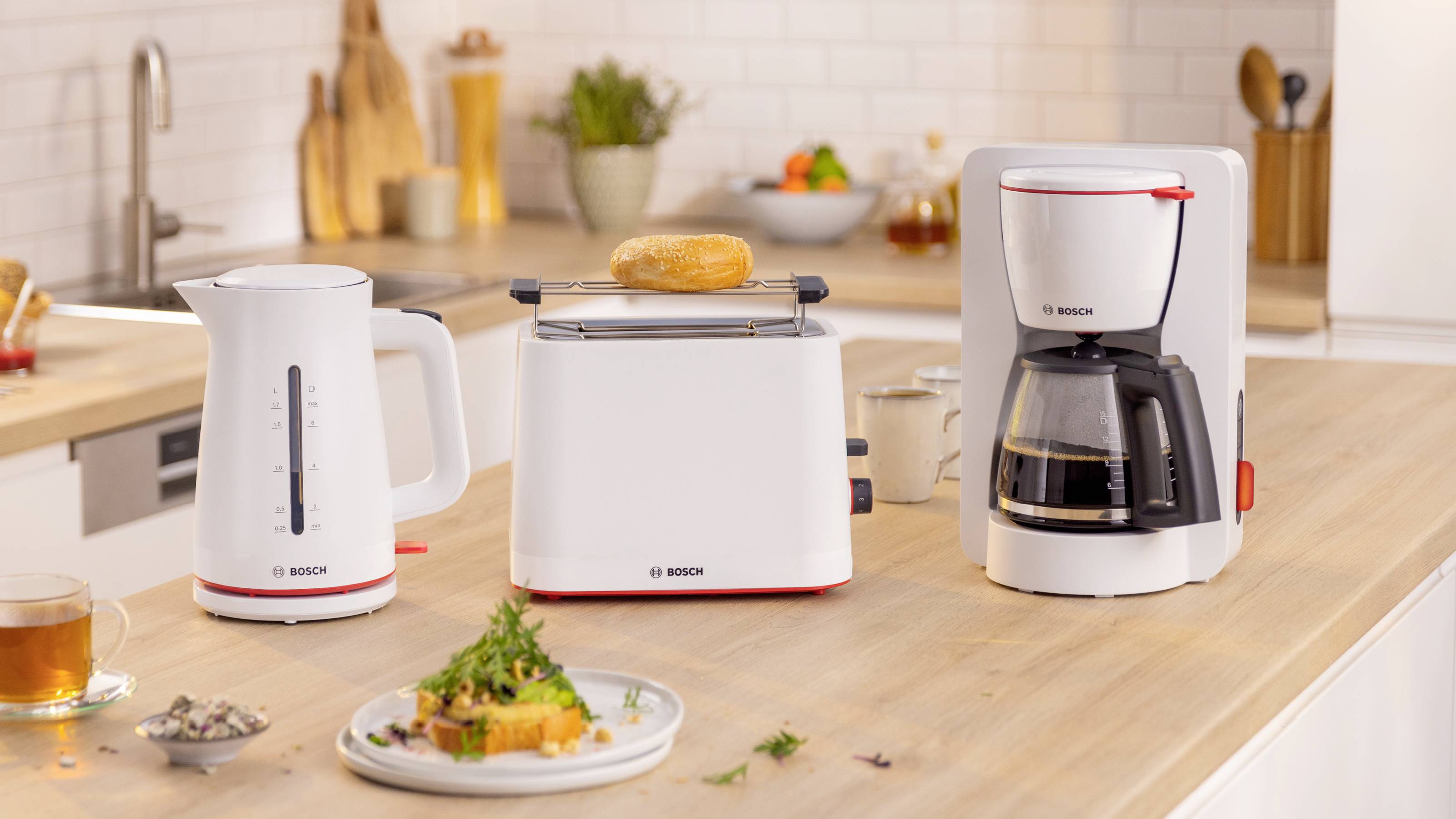 A modern kitchen setting with a white kettle, toaster with a bagel, and coffee maker on a wooden counter. Plates with food and a tea cup are nearby.