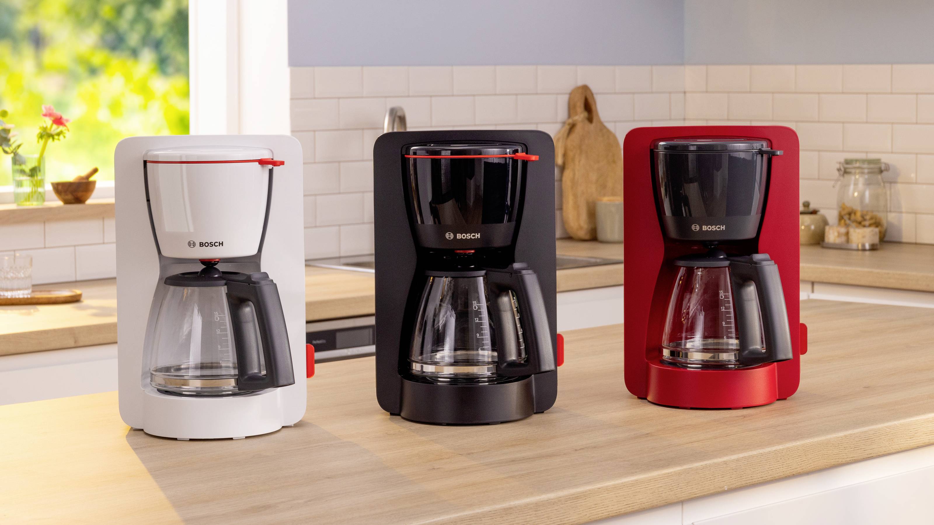Three Bosch coffee makers in white, black, and red are displayed on a kitchen counter with a tiled backsplash and bright window view.
