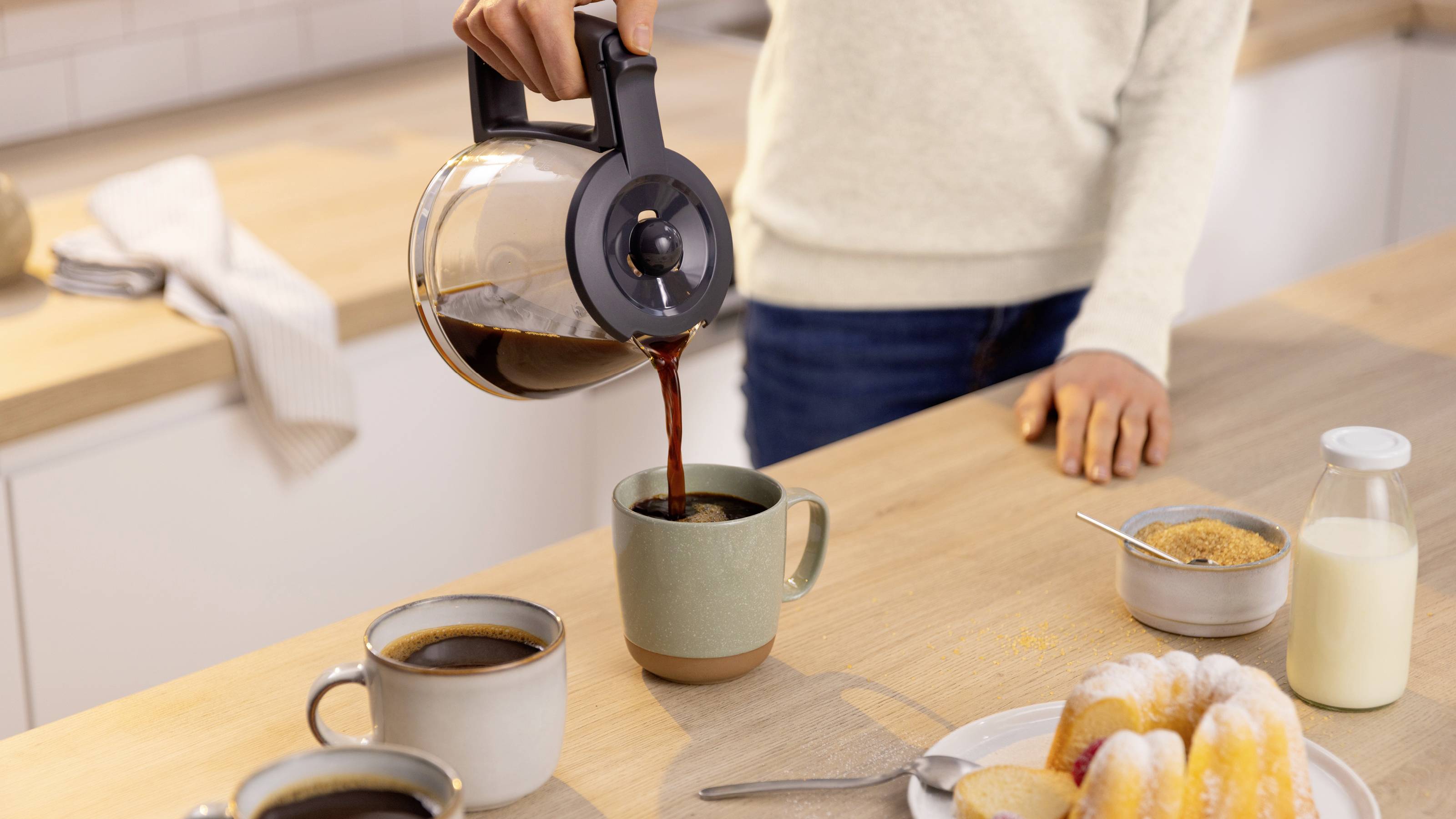 A person pours coffee into a mug on a kitchen counter. Nearby are two filled cups, a plate of pastries, a jar of sugar, and a milk bottle.