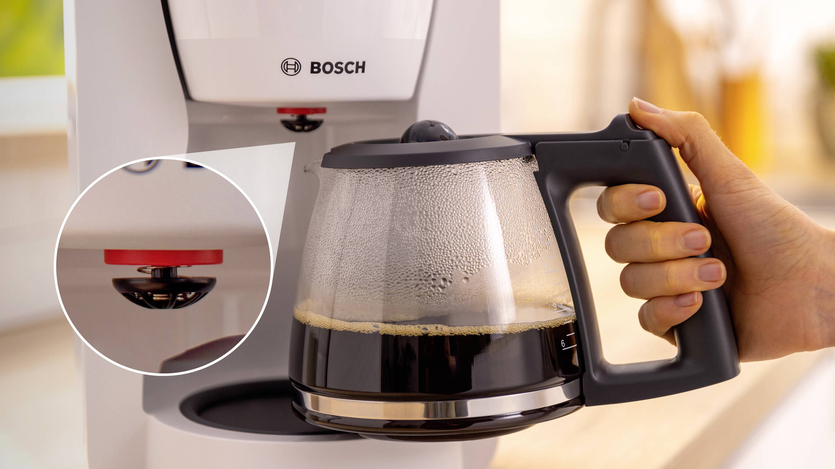 A hand holds a coffee pot filled with freshly brewed coffee being placed into a Bosch coffee maker. A red button detail is highlighted.