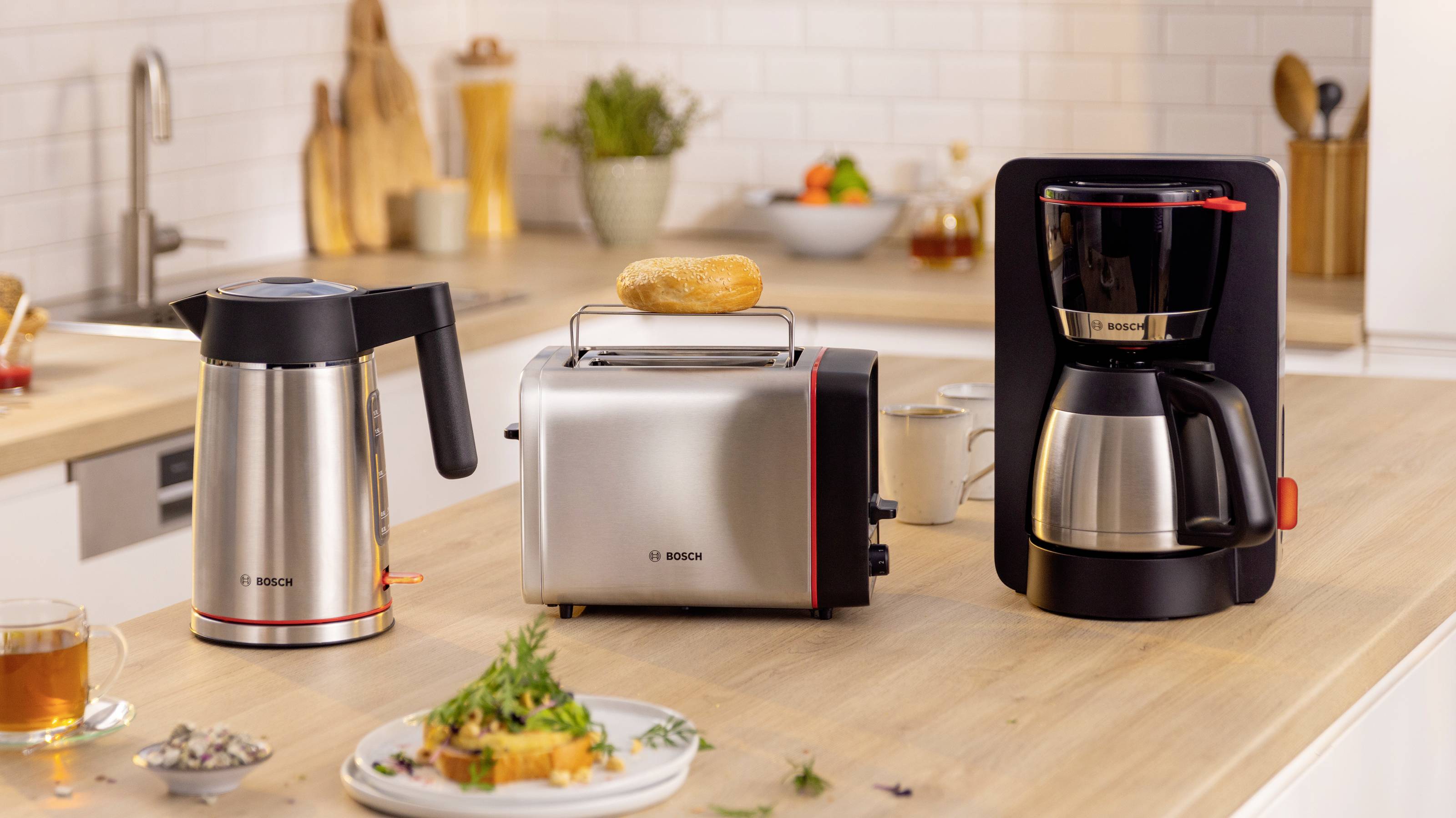 A modern kitchen counter displays a silver coffee maker, toaster with a bagel, and an electric kettle. Fresh ingredients and a cup of tea are nearby.