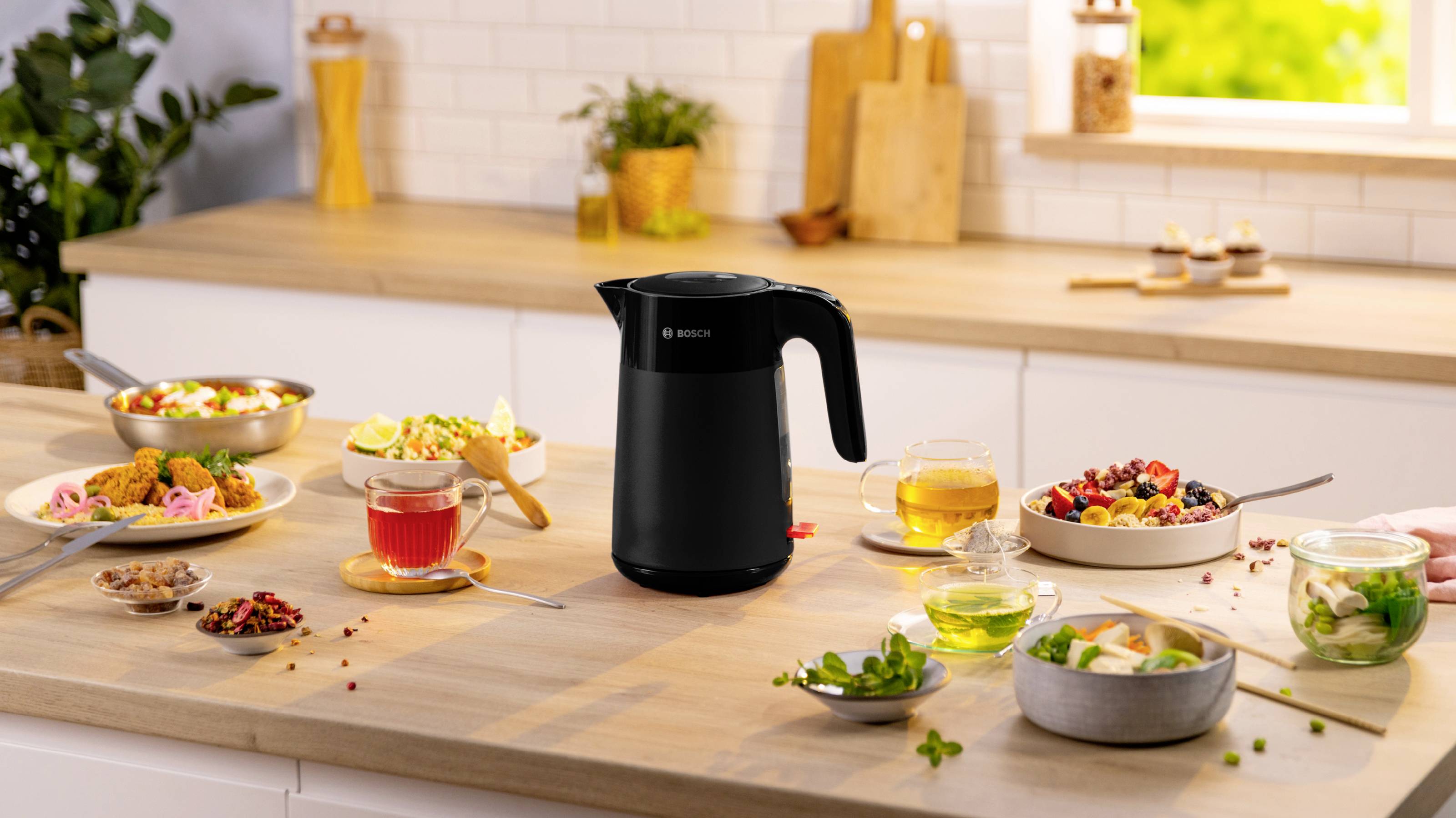 A black electric kettle sits on a kitchen counter surrounded by bowls and cups of colorful salads, tea, and ingredients, set against a bright kitchen backdrop.