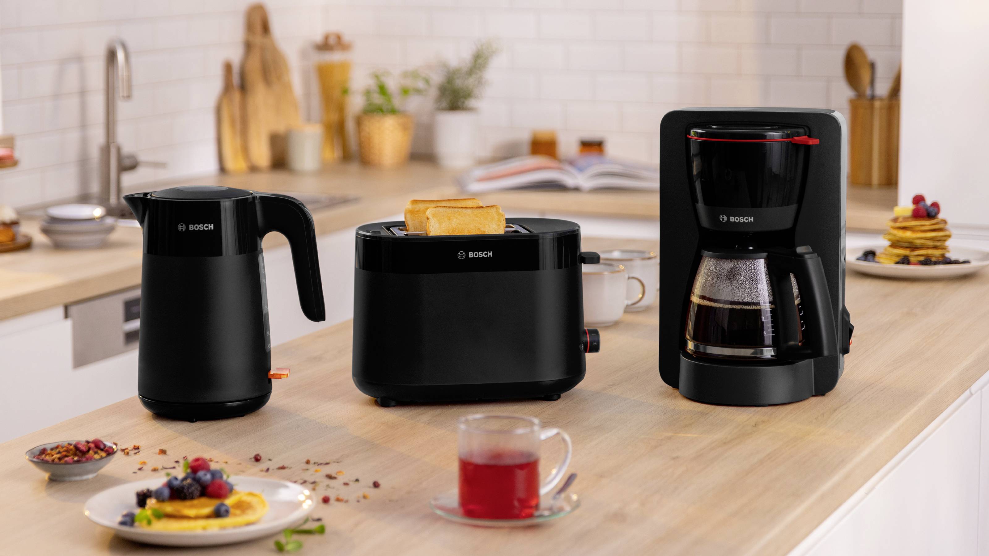 A modern kitchen countertop displaying a black kettle, toaster with toast, and coffee maker. In the background, pancakes and fruit are visible.