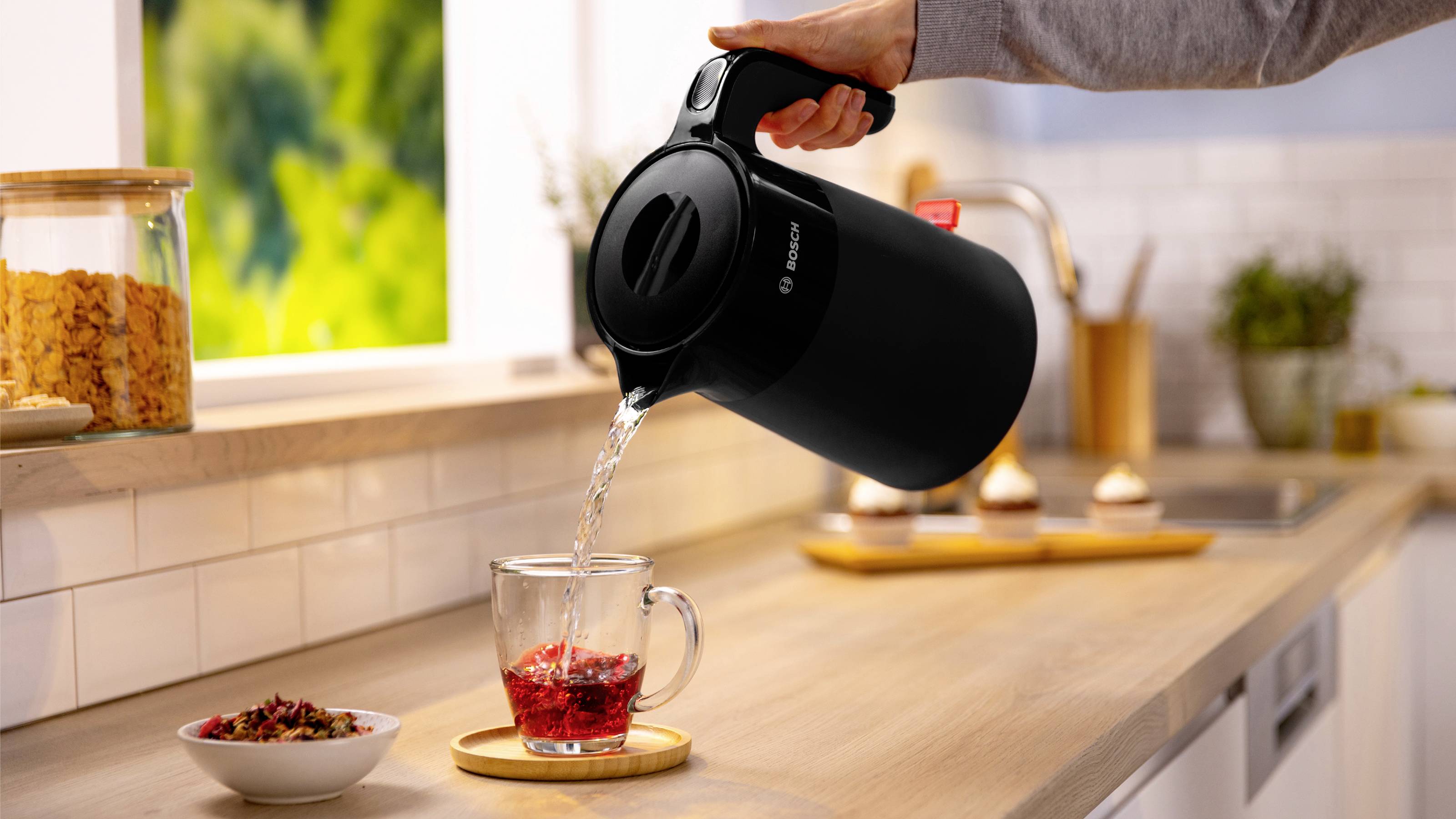 A person pours hot water from a kettle into a glass cup with herbs on a kitchen counter, next to a window and a jar of cereal.