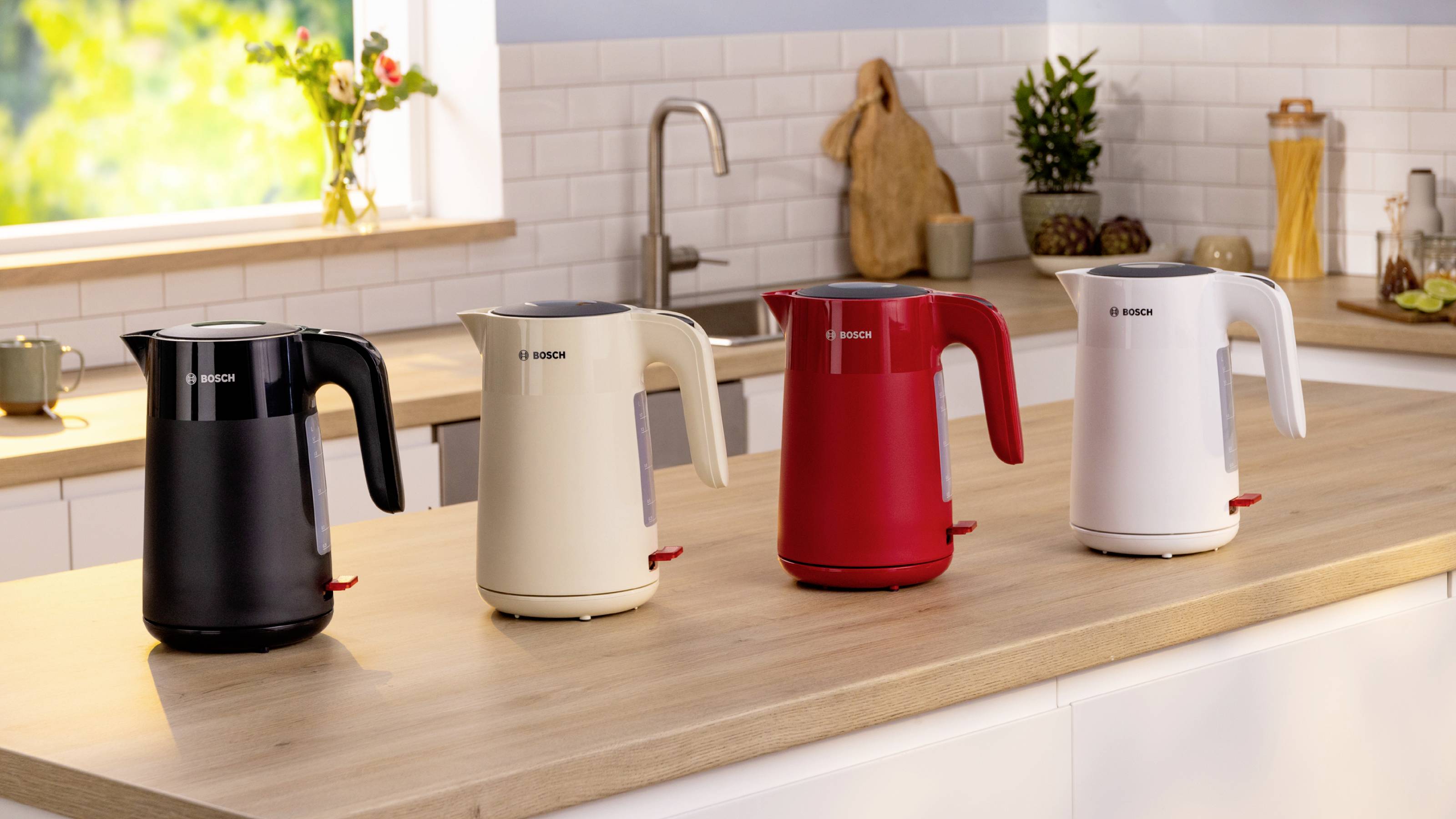 Four electric kettles in black, beige, red, and white are aligned on a kitchen counter with a blurred, sunny background.