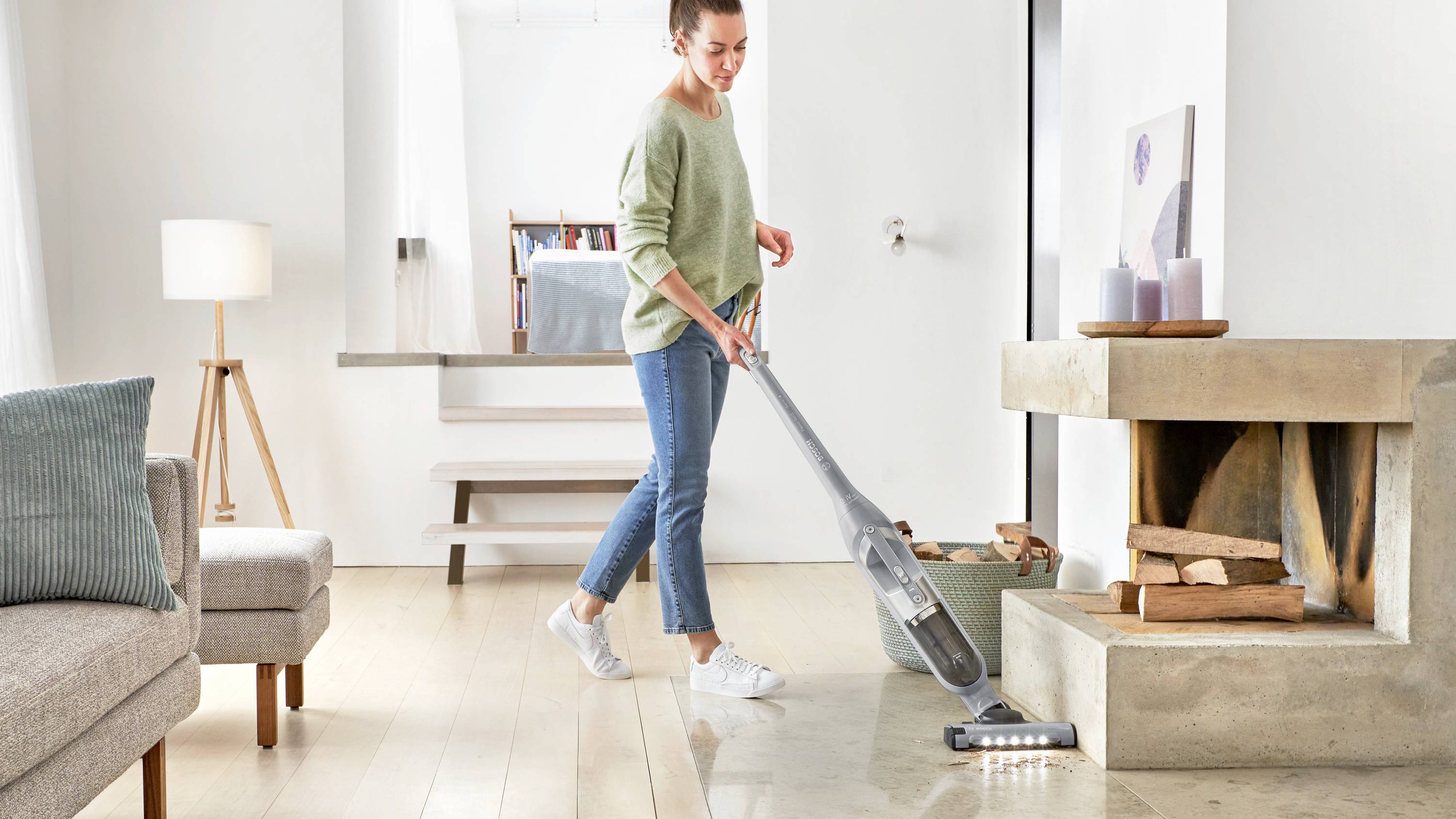 A person vacuums a modern living room with a cordless vacuum, featuring a sofa, lamp, and a fireplace with stacked wood in the background.