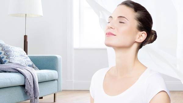 A woman in a white shirt sits peacefully, eyes closed, in a bright room with a sofa and lamp in the background.