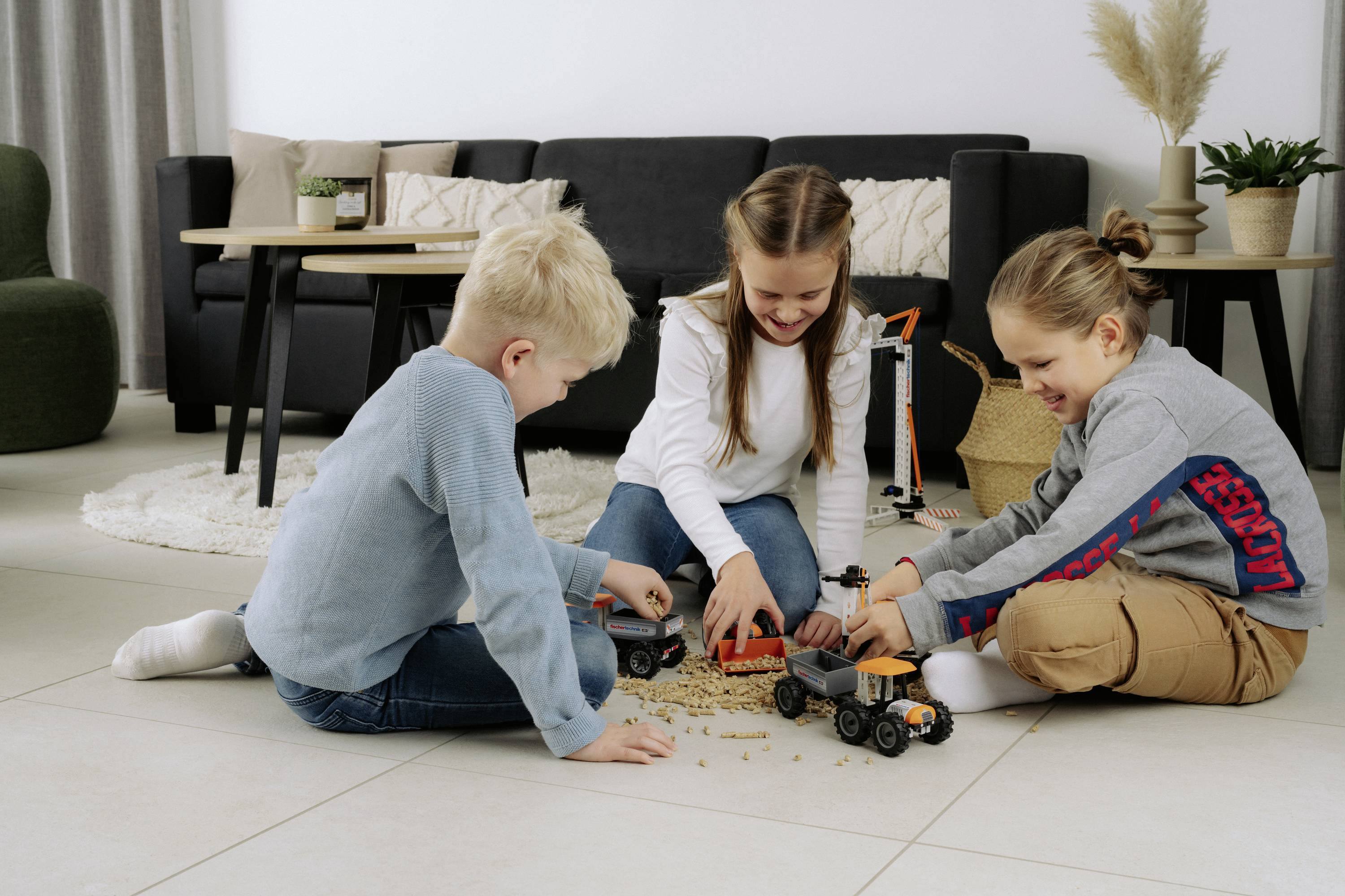 Three children are playing with toy trucks on the floor in a living room, smiling and interacting with each other, creating a playful scene.