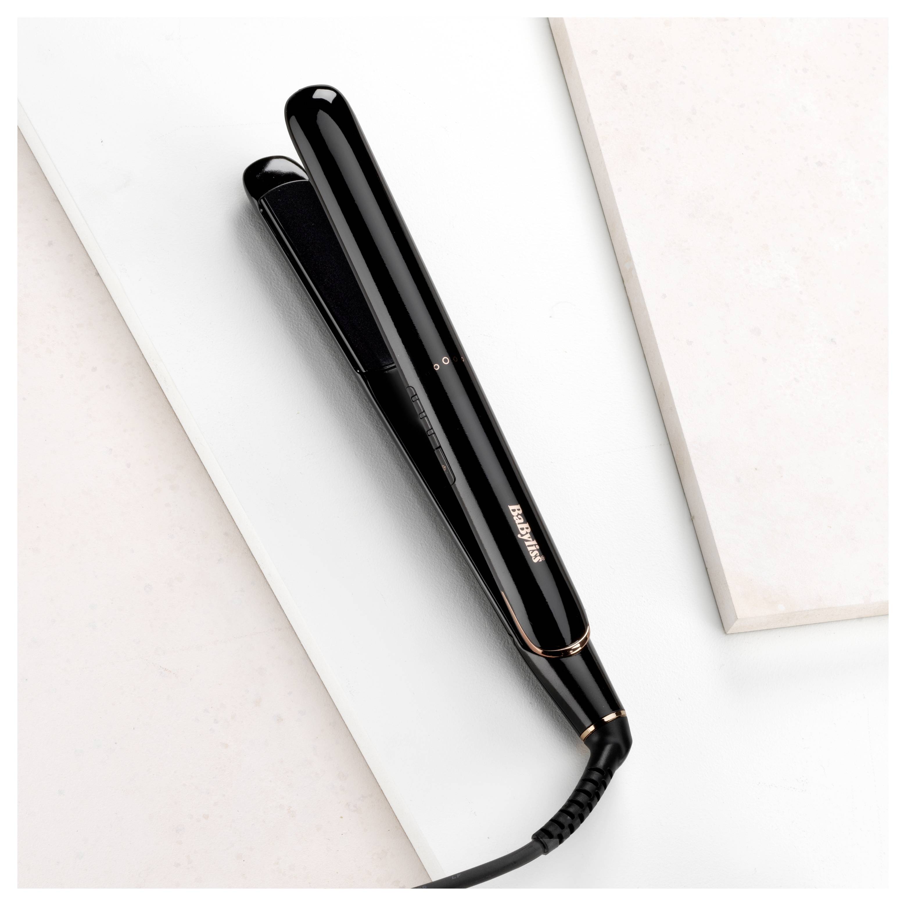 A black hair straightener with a glossy finish is placed on a white marble surface next to a beige tile, showcasing a sleek design.