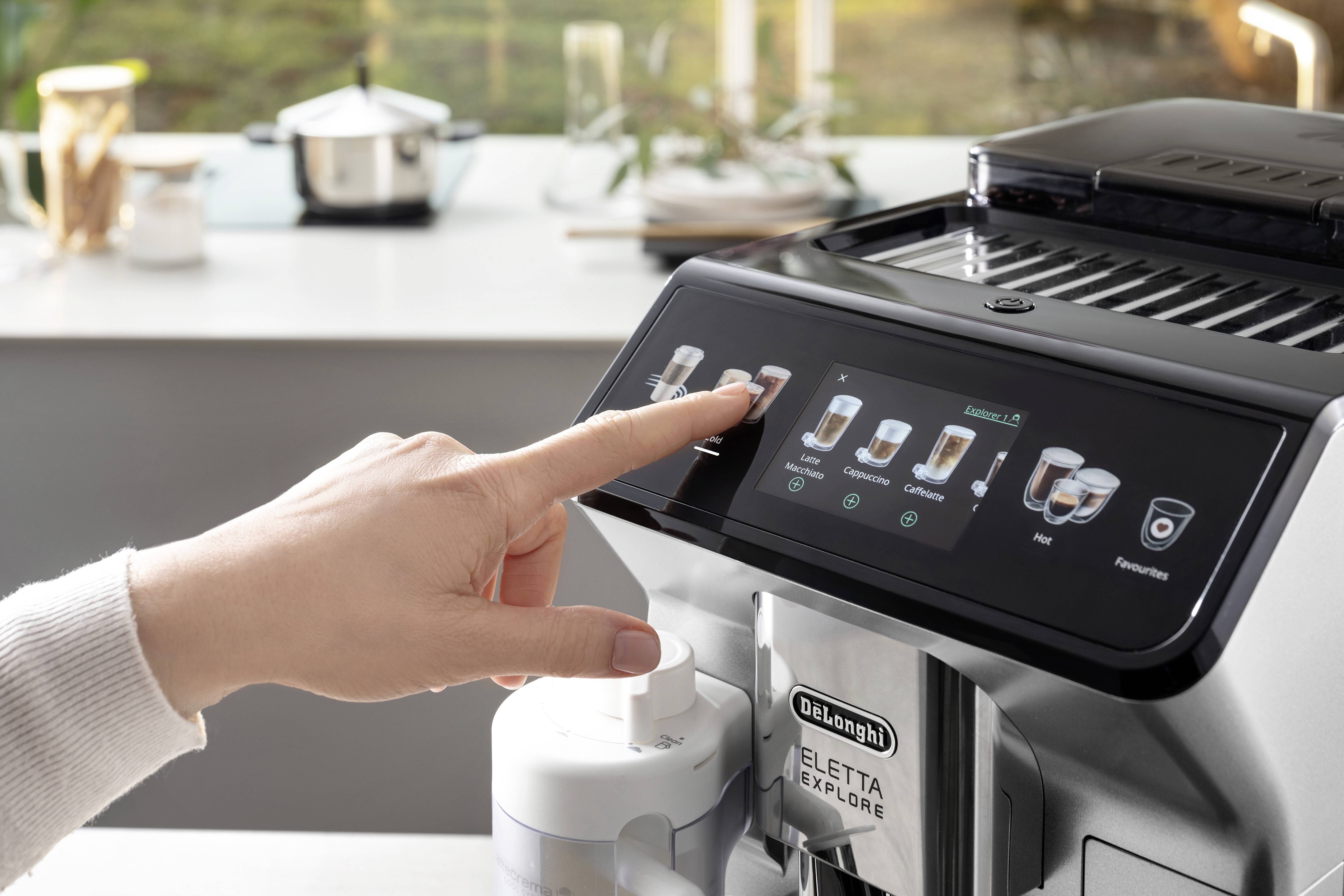 A hand selects a drink option on the touchscreen of a De'Longhi Eletta Explore coffee machine in a modern kitchen setting.