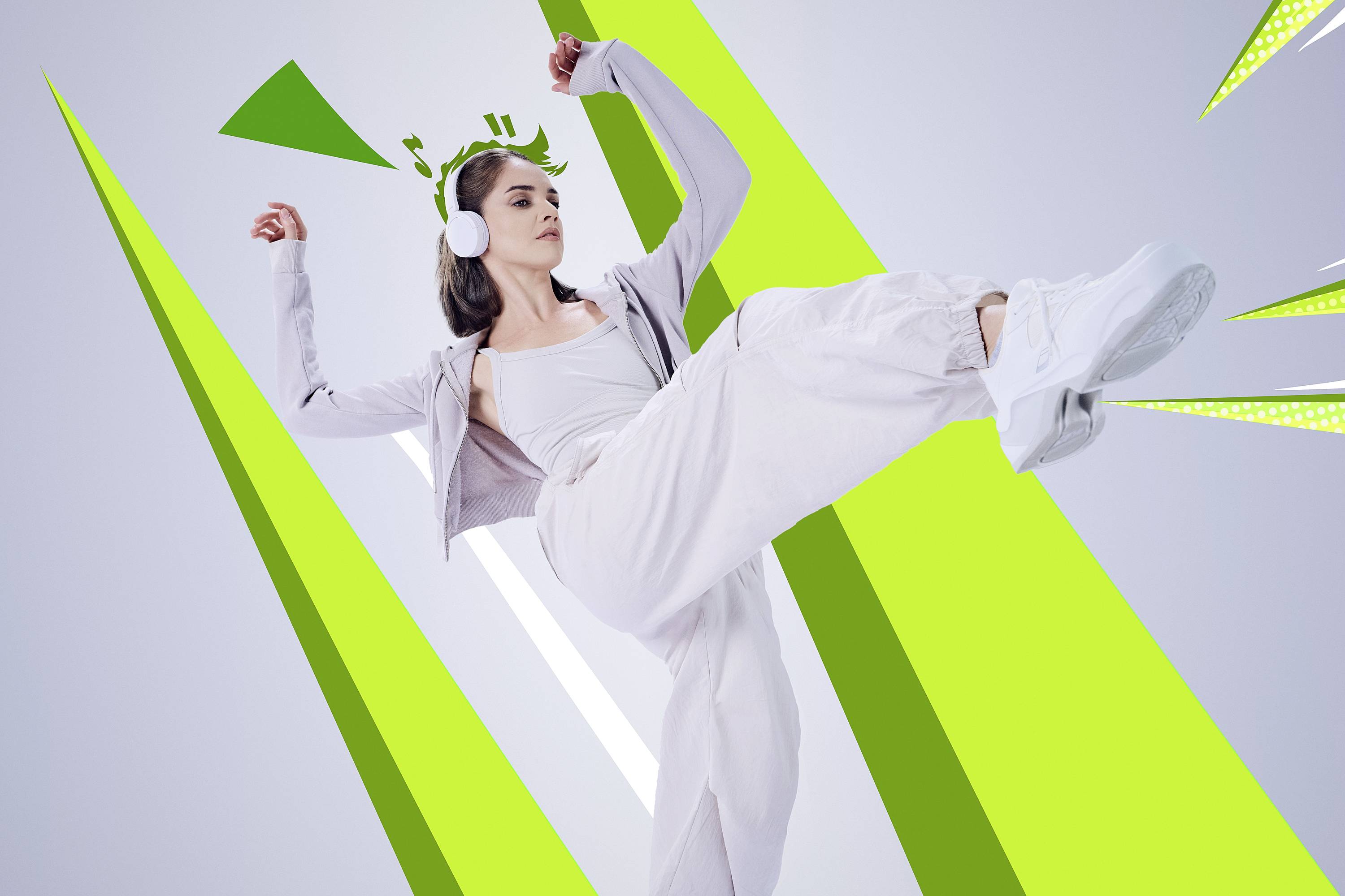 A person wearing headphones and casual clothing performs a high kick against a background of dynamic green and white geometric shapes.