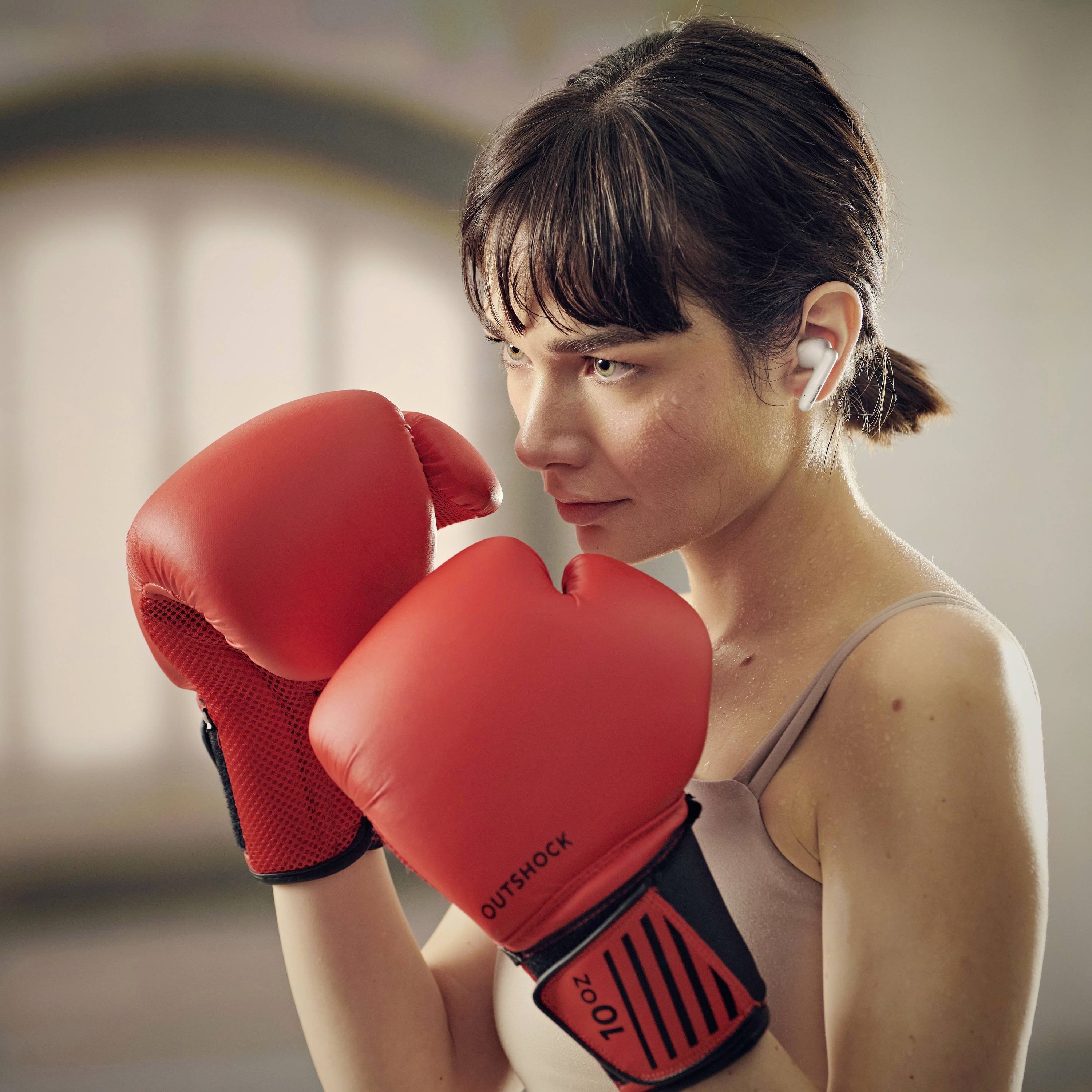 A woman wearing boxing gloves stands in a fighting stance, focusing intently. She wears wireless earbuds, suggesting fitness or training.