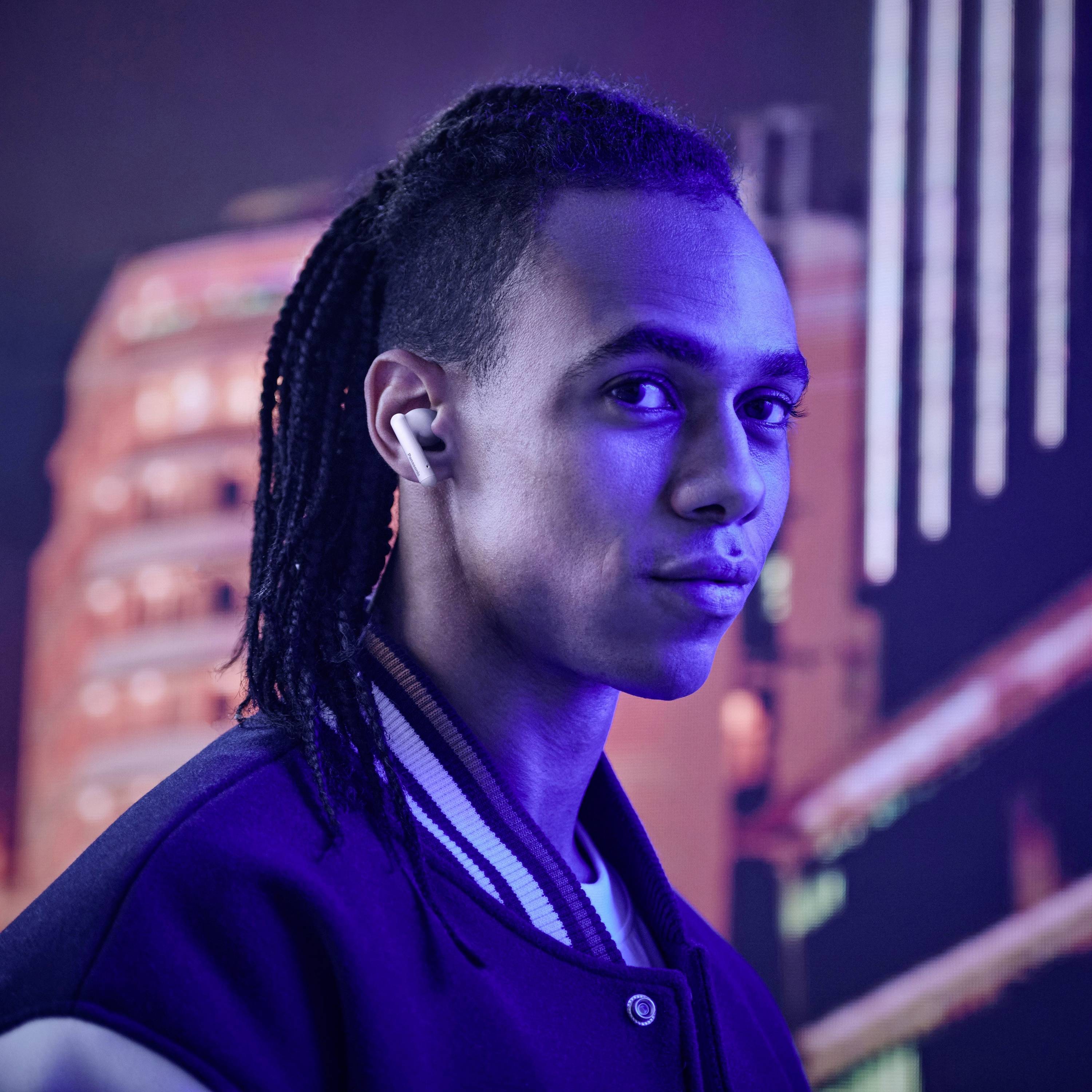 A person wearing a varsity jacket and earbuds looks over their shoulder in a cityscape at night, with colorful neon lights in the background.