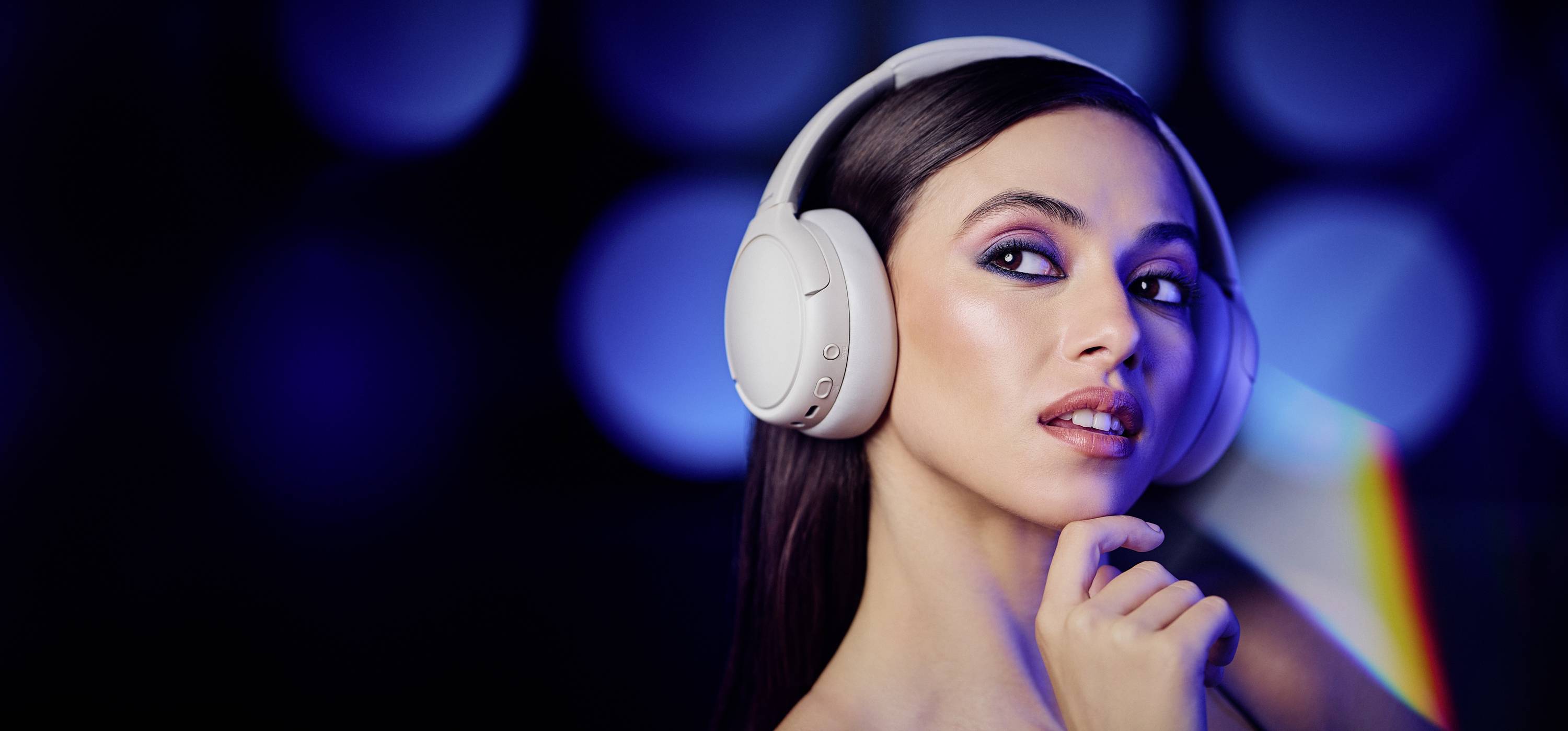 A woman wearing white headphones stands against a blurred, multicolored background. She looks over her shoulder with a contemplative expression.