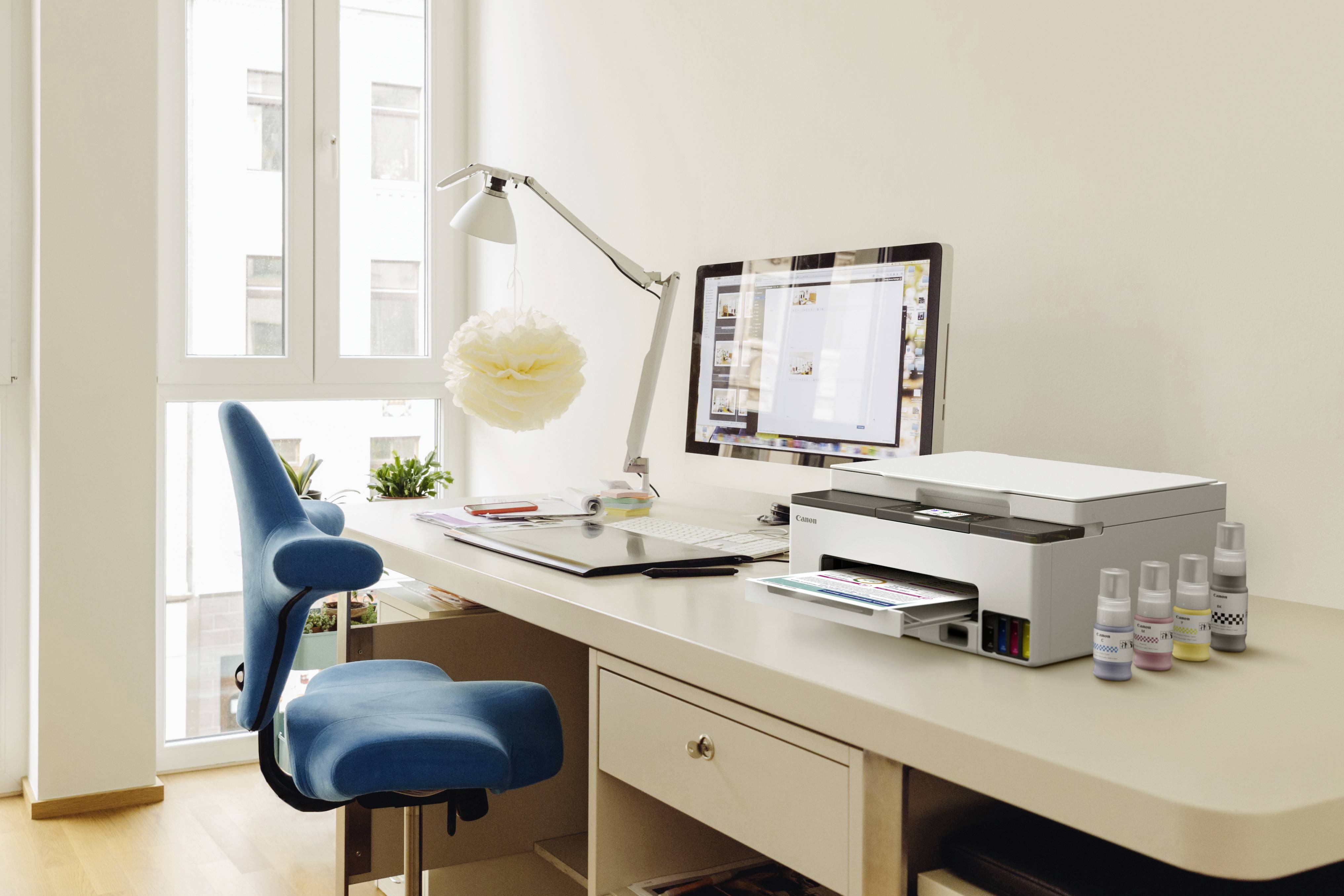 A bright home office features a modern computer, a printer on a desk with office supplies, a blue chair, and a large window with natural light.