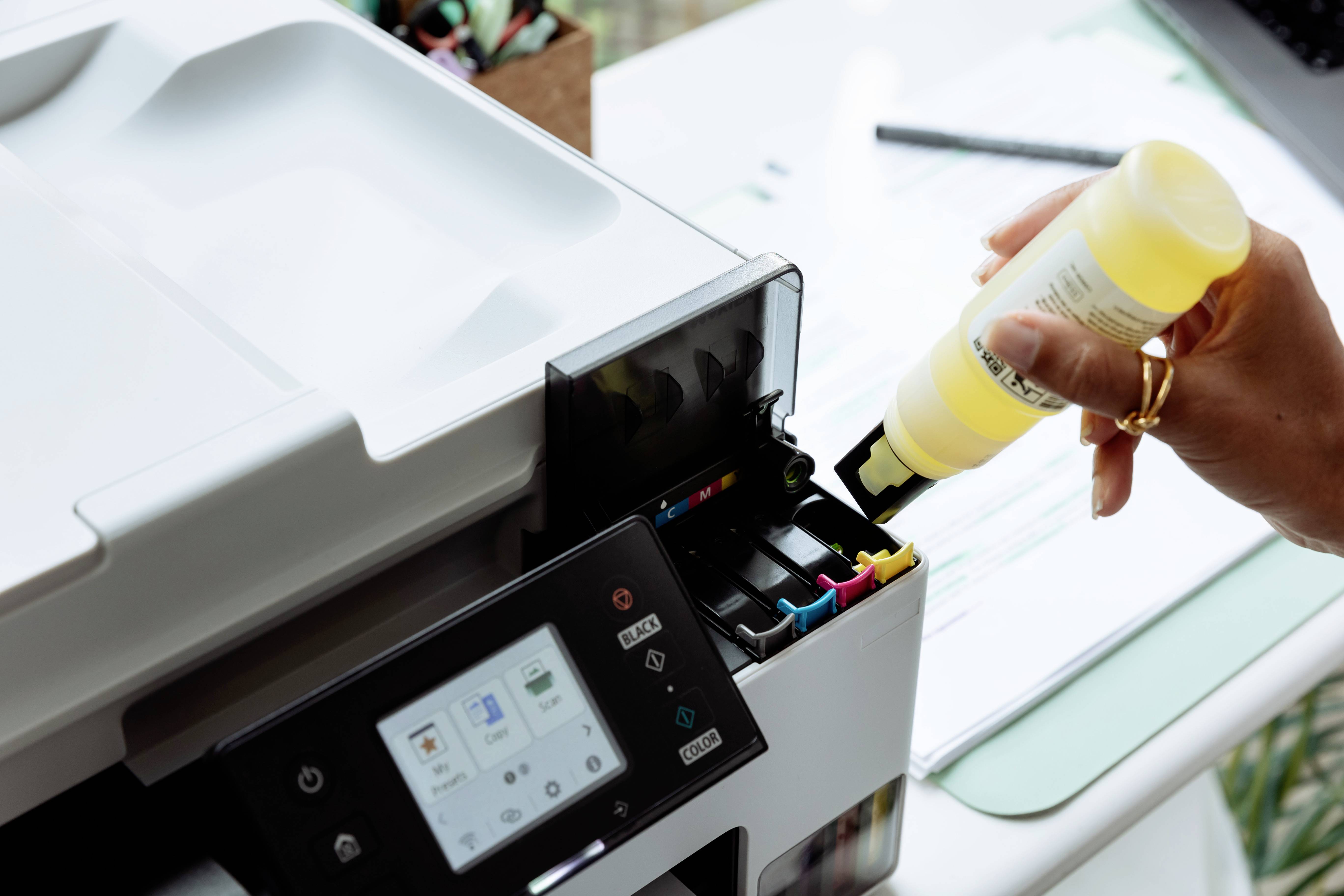 A person refills an ink printer with yellow ink, showing the printer's display and ink cartridge area.